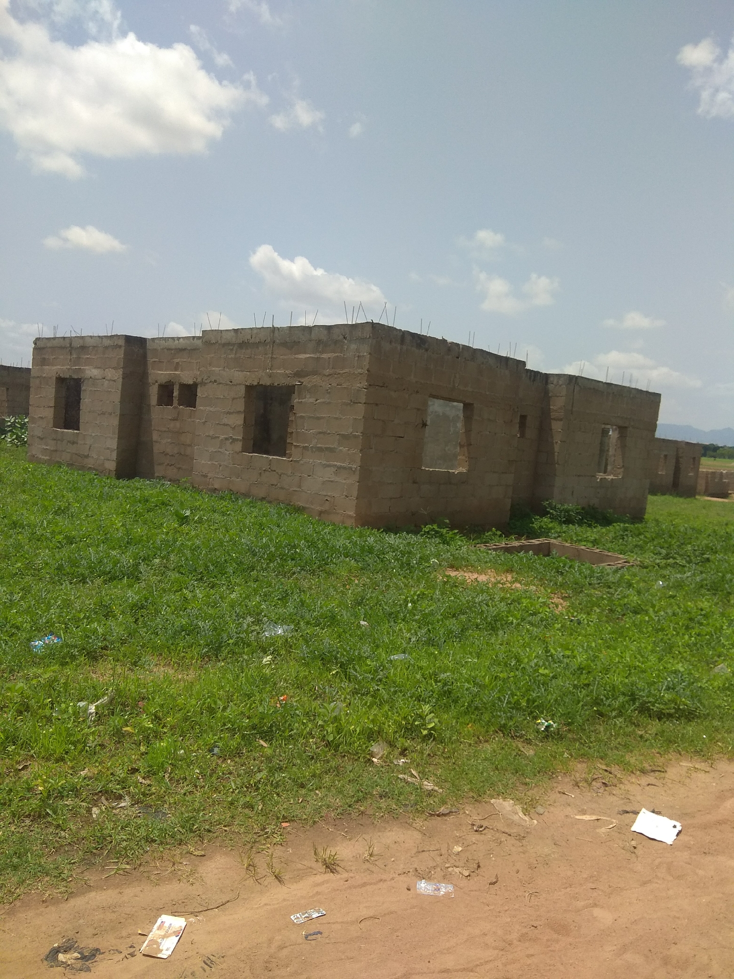 The image shows a construction site in Jalingo, Nigeria, where several unfinished buildings are under construction. The buildings are made of concrete blocks, with exposed rebar at the rooflines, indicating that the structures are in the early stages of completion. The buildings have open doorways and window frames, with no glass or doors installed. The ground is covered with green grass, which appears to be overgrown in some areas, and there is some litter scattered on the ground. The sky is partly cloudy, with a few scattered white clouds against a pale blue sky. In the background, there are distant hills or mountains. The foreground of the image shows a dirt path with some litter, including what appears to be paper and plastic. The overall impression is of a developing area with ongoing construction projects.