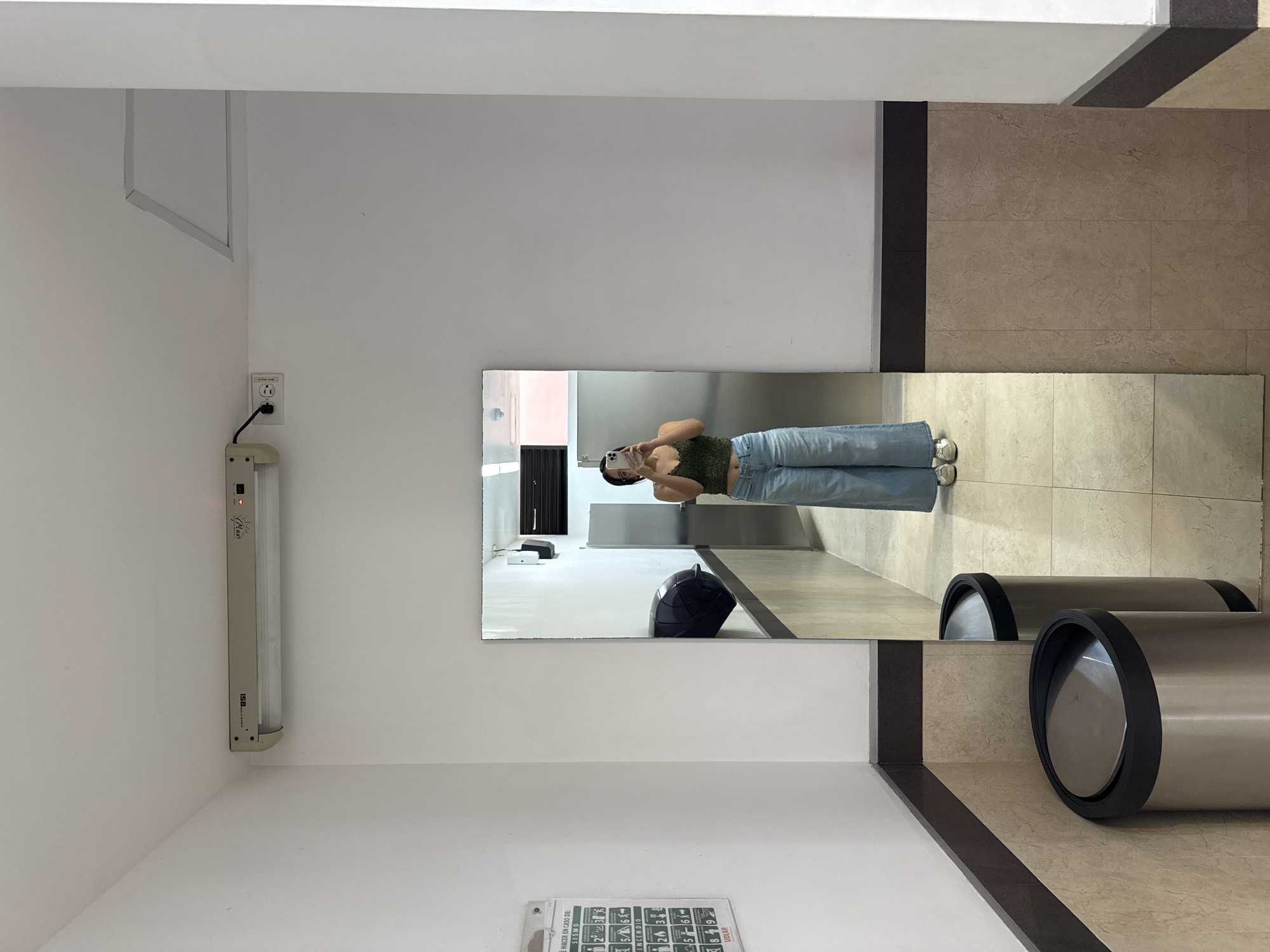 An indoor, well-lit scene in San Andrés Cholula, Mexico, depicts a person taking a mirror selfie. The image is oriented vertically, showing a white-walled room with light-colored, possibly beige, tiled flooring accented by darker brown strips.
The central subject is a person, likely female, reflected in a large, full-length mirror. They are wearing a dark green, possibly lace or textured, crop top and light blue, wide-leg jeans, along with light-colored shoes. The person holds a white smartphone at arm's length, angled towards the mirror to capture their reflection.
To the left of the mirror, mounted on the white wall, is a horizontal fluorescent light fixture. It has text visible, including "Maxi" and "ISB," and a small red indicator light. Below the light fixture, an electrical outlet with a plugged-in cord is visible.
In the foreground, outside the mirror's reflection, are two cylindrical, metallic trash cans with black lids or rims, situated on the tiled floor near the base of the wall. These same trash cans, or similar ones, are also partially visible as reflections within the mirror, positioned behind where the camera would be. Another dark, rounded object, possibly a trash receptacle, is also visible on the floor within the mirror's reflection, further back.
On the lower left section of the wall, a piece of paper or sign with text in Spanish is affixed. Portions of the text read "HACER EN CASO DE," "SISMO," and "INCENDIO," along with numbered steps, suggesting it is an emergency procedure or instruction sheet. The overall setting appears to be a public or semi-public space, such as a restroom, hallway, or changing area.