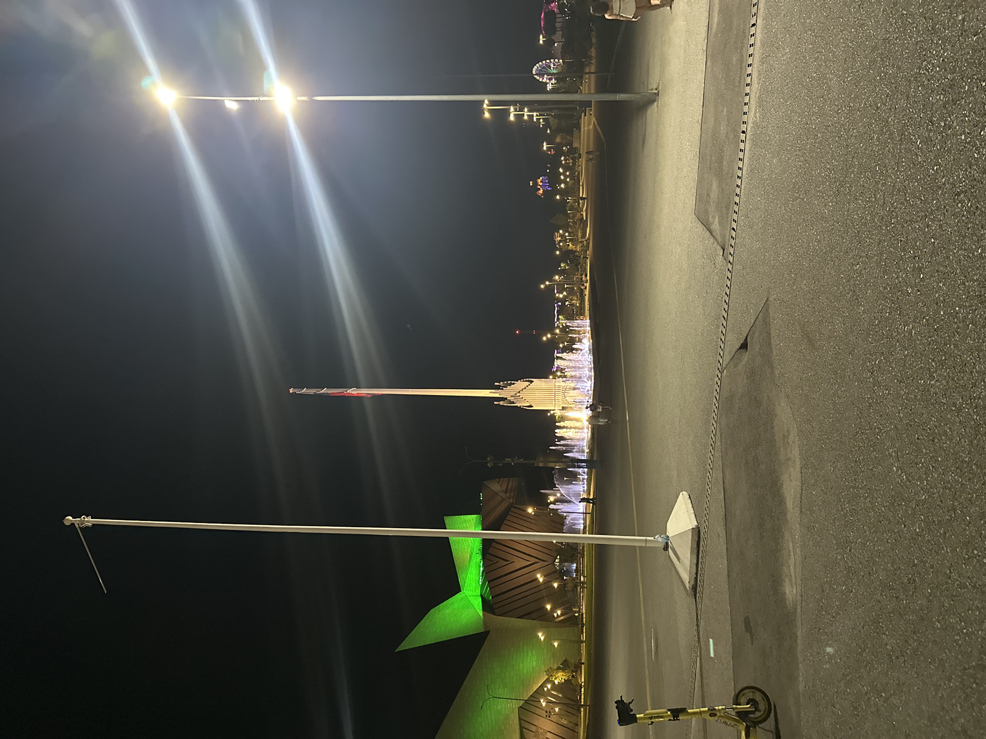 At night in the city of Sirius, Russia, a wide, paved area is illuminated by streetlights. In the foreground, a yellow electric scooter is parked on the left. Further back, a tall white flagpole stands with a diagonal spar extending from its top. Beyond the flagpole, a large, modern building with a distinctive green, geometric facade is lit up. In the mid-ground, a fountain displays illuminated water jets. A tall, ornate structure, possibly a monument or part of the building complex, stands near the fountain. In the background, a Ferris wheel and other amusement park rides are visible, along with numerous streetlights and illuminated buildings, suggesting a recreational area. A lone female figure is walking away from the camera on the right side of the frame. The sky is dark, indicating nighttime.