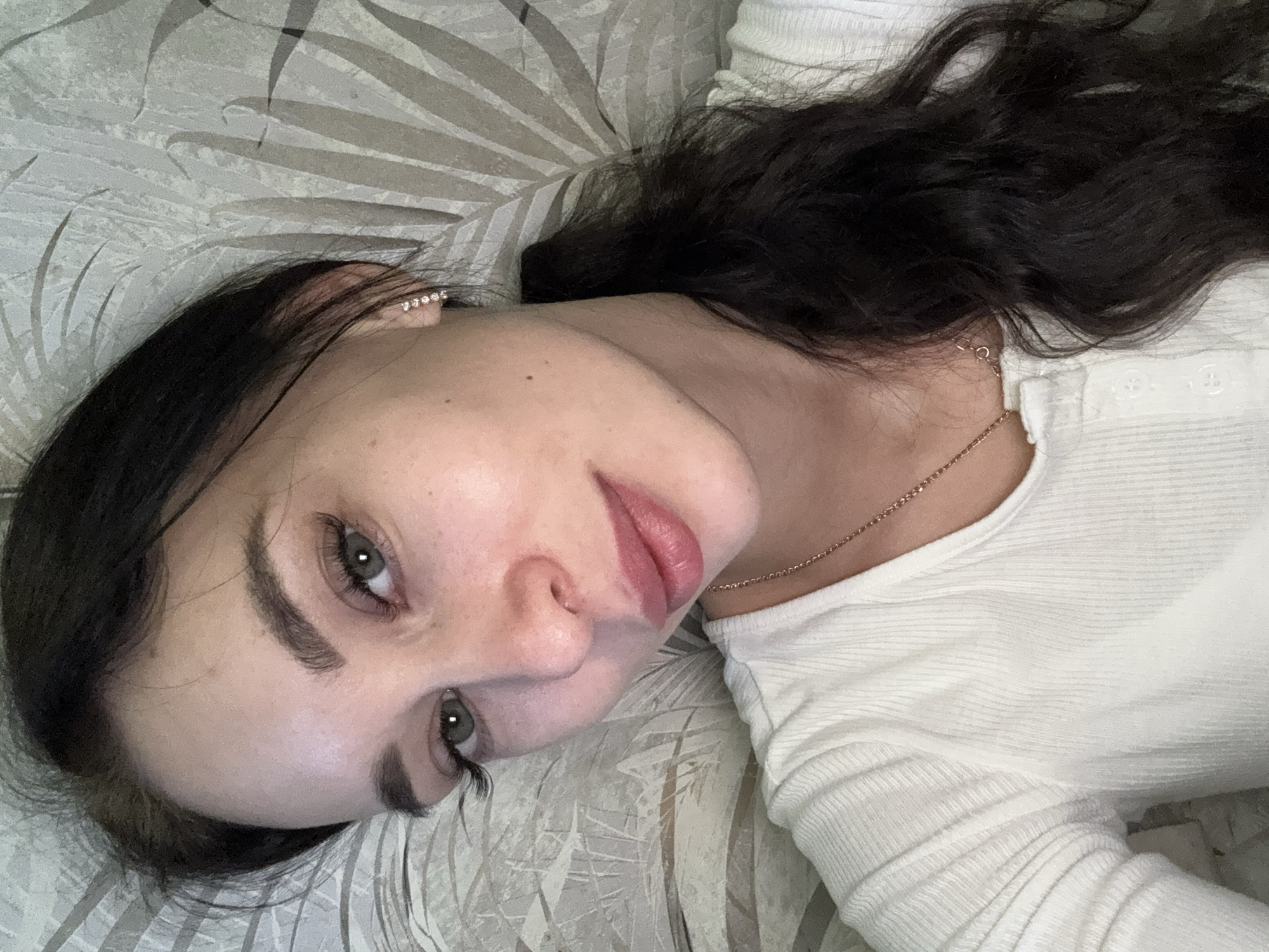 A woman with dark hair and light eyes is lying on a patterned surface. She is wearing a white long-sleeved shirt and a delicate gold necklace. She also has earrings. The location is unknown, but it is implied to be in Chaikovsky, Russia.