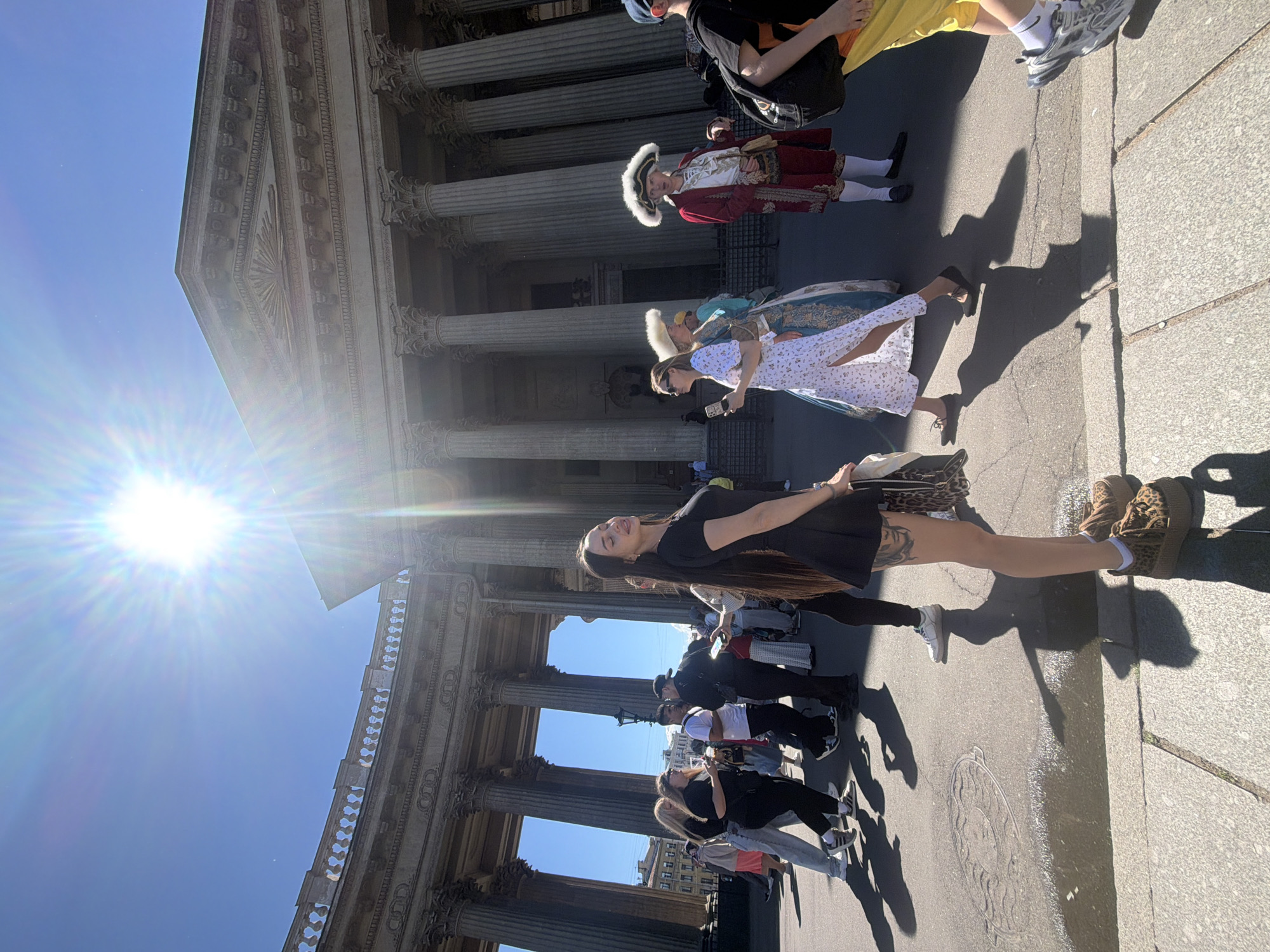 The image shows a sunny day in front of the Kazan Cathedral in Saint Petersburg, Russia. A woman in a black dress and leopard-print shoes is standing in the foreground, looking towards the sun. She has long, dark hair and a tattoo on her leg. Behind her, a group of tourists and locals are walking around, some dressed in modern clothing, others in historical costumes. The Kazan Cathedral, with its impressive colonnade, is visible in the background. The sun is shining brightly, casting long shadows on the ground.
