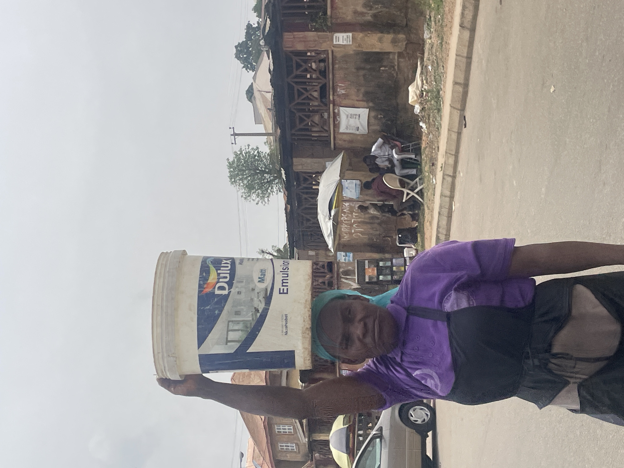 A woman carries a large bucket of Dulux Matt Emulsion paint on her head while walking along a street in Jos, Nigeria. She is wearing a purple shirt, black apron, and head covering. A car is partially visible next to her. In the background, there is a building, possibly a market or shop, where people are seated, and a street with vegetation and other buildings. The sky is overcast.