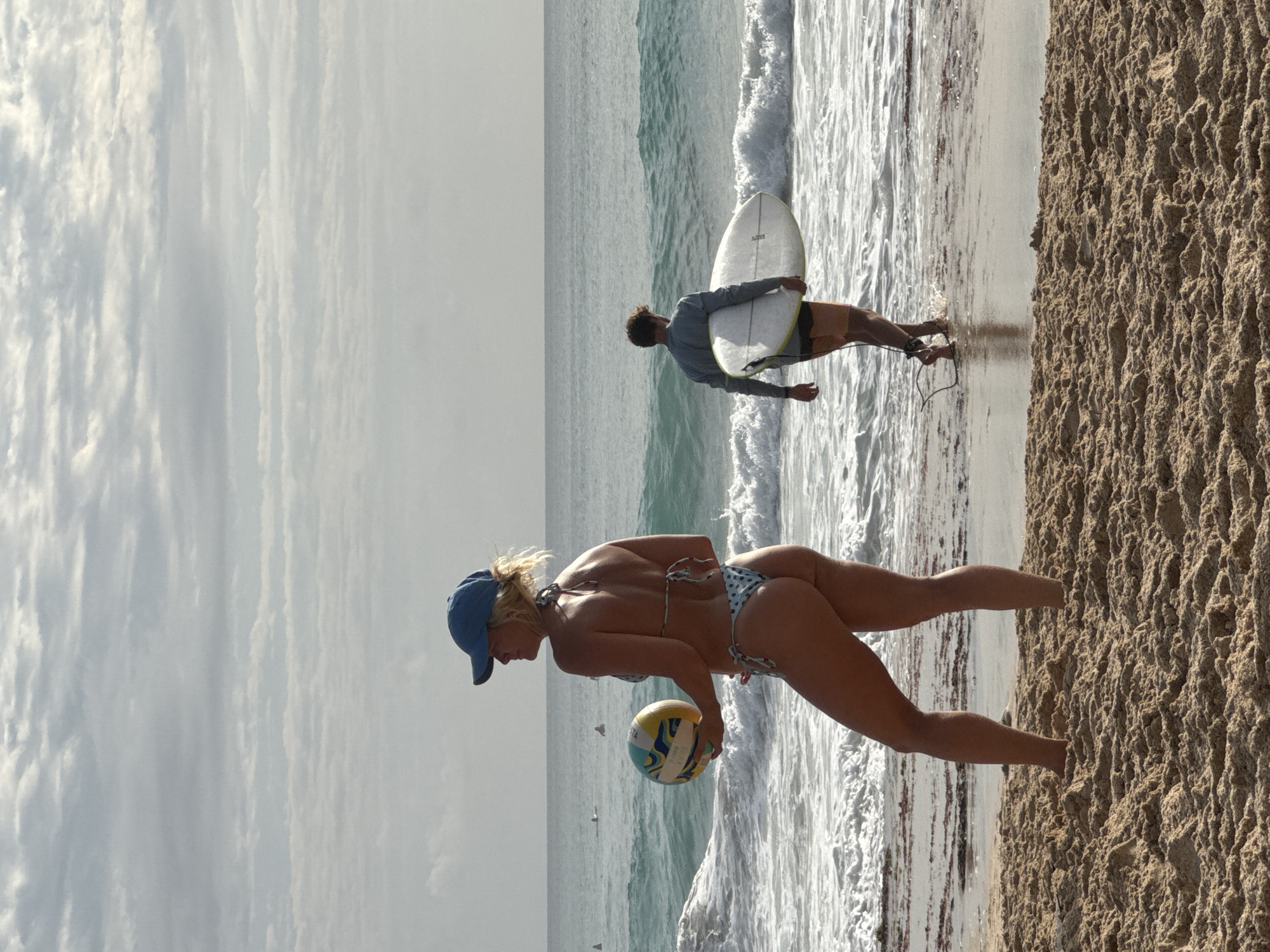 This is a vertical, full-frame, eye-level shot of a beach scene in Kuta Selatan, Indonesia. In the foreground, a woman wearing a blue baseball cap and a patterned bikini stands on the sandy beach, holding a colorful volleyball. Her body is angled to the left, and she is looking out at the ocean. Behind her, the ocean is a vibrant turquoise, with waves breaking and white foam rolling onto the shore. In the mid-ground, a man with short brown hair, wearing a long-sleeved grey top and orange shorts, walks into the ocean carrying a white surfboard. The ocean stretches to the horizon, where it meets a pale, cloudy sky. There are a few distant figures in the water, possibly other surfers. The overall lighting suggests it is either early morning or late afternoon, with soft, diffused light. The sand in the foreground is textured with small pebbles and ripples.