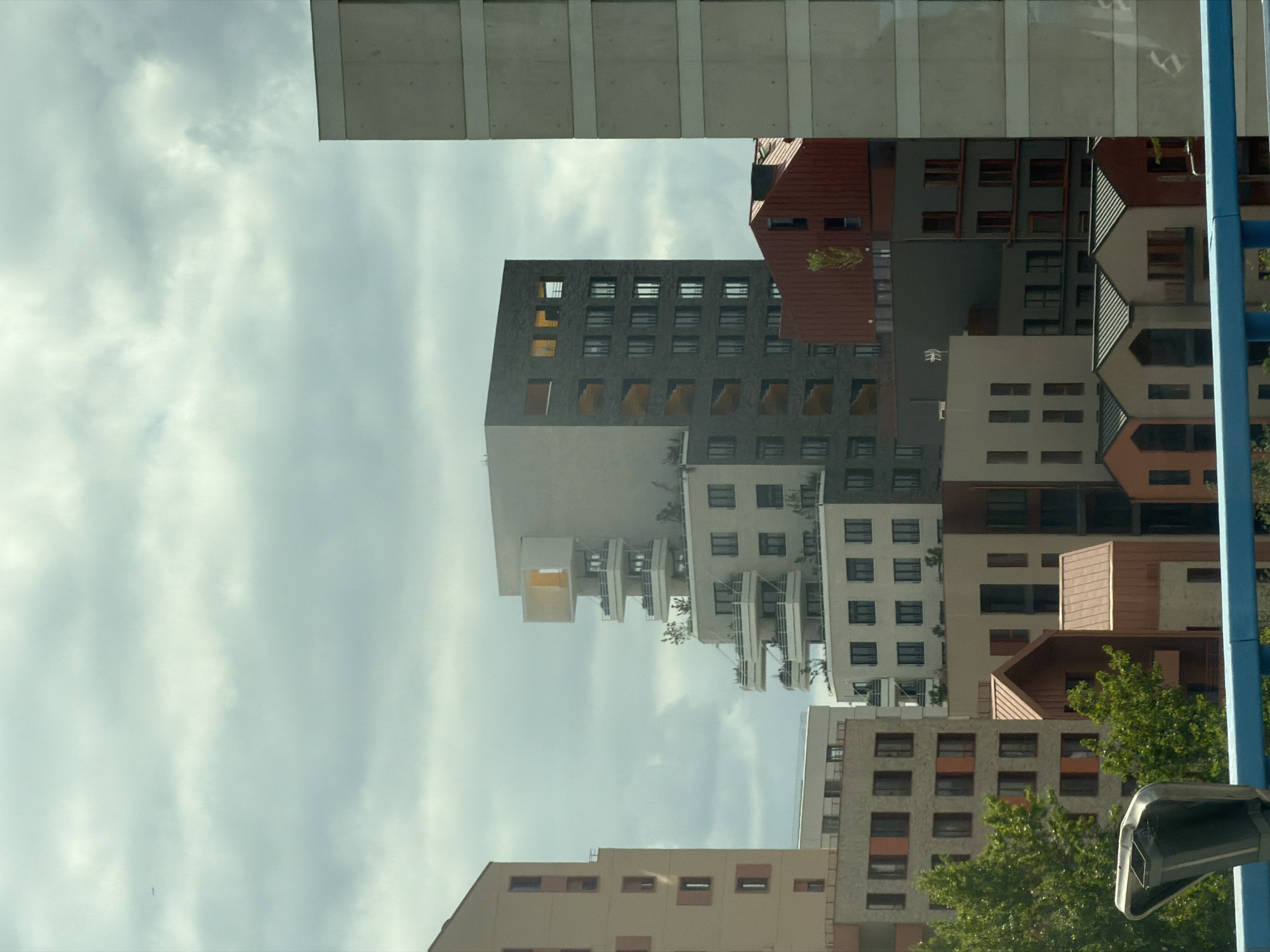 This is a vertical, low-angle photograph of a cluster of modern apartment buildings against a cloudy sky. The buildings are of varying heights and architectural styles, with many featuring balconies and dark-framed windows. Some buildings have red-tiled roofs and brown or grey facades, while others are lighter in color. Green trees are visible in the lower right portion of the image, suggesting a residential or urban park setting. The sky is overcast with soft, diffused light, indicating it could be daytime, possibly late afternoon or early morning, or simply a cloudy day. There is a bright blue pole on the right side of the image, and part of a black security camera is visible in the bottom right corner. The image appears to be a reflection on a window or glass surface, as indicated by a faint reflection of what looks like a ceiling or internal structure at the top of the image. The scene is quiet and appears to depict an urban landscape in Alfortville, France. There is no visible text in the image.