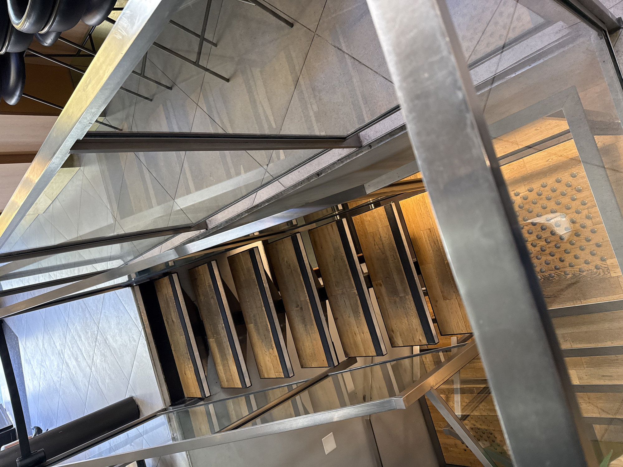 The image shows a downward view of a modern staircase, likely inside a building. The steps are constructed with wood and dark accents, possibly metal. Glass panels serve as railings, secured by metal frames. The flooring surrounding the staircase appears to be tile and wood. There are no people visible in the image. The location of the image within the city of Paris, France is not discernable from the image.