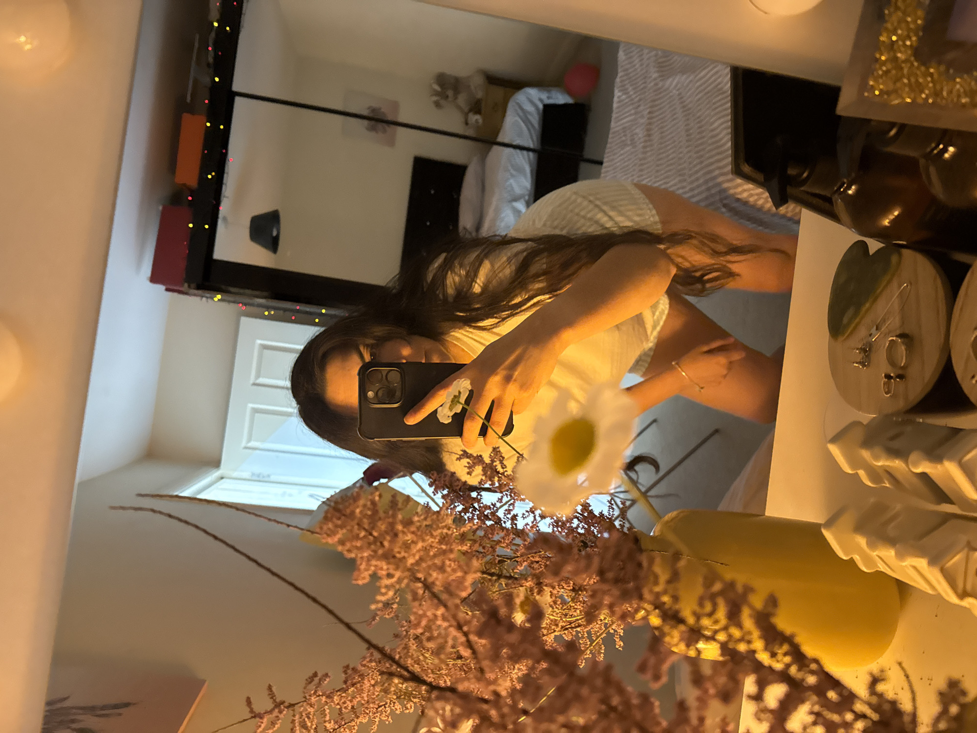 A person is taking a selfie in a mirror. The mirror shows a bedroom in the background, featuring a bed and a wardrobe with decorative lights. The foreground of the mirror's reflection is obstructed by flowers and a yellow bottle. The person is holding a phone up, capturing the image. The location is not specified but the city is stated as Great Yarmouth, United Kingdom.