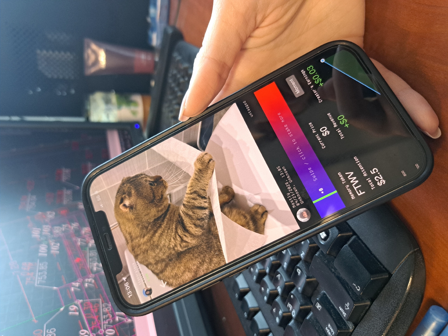 An overhead shot captures a hand holding a black smartphone over a computer keyboard and monitor in what appears to be a workspace or home office.
The primary subject is a dark-cased smartphone held at an angle by a person's hand, revealing its screen content. The screen prominently displays an image of a brown tabby cat with bright green eyes, seen from the side, stretching with one paw raised. Above the cat's image, the phone's status bar shows "13:06" and "5G". Below the cat, text includes a profile picture next to "georgieve", followed by "Nestly", "04/21/2026, 13:05", and "Unknown, Unknown". Further down, a prompt reads "Swipe / click to stake more" and "untagged". On the right side of the screen, various metrics are displayed: "Memory Token", "FTWV", "Total Attention $2.5", "Current Price $0", "Total Revenue +$0", and "Creator's Earnings +$0.03". A colorful bar (red, blue, green segments) and a "Moment" button are also visible.
In the background, an ergonomic black computer keyboard is partially visible on a wooden desk surface. Behind the keyboard, a computer monitor displays complex graphs and numbers, indicative of financial or stock market data, including "753.85", "100", "303", "19", "74", and "0753.82". Other blurry objects, possibly bottles or containers, are also present in the background.
The activity depicted is a person viewing content on a smartphone, possibly interacting with a social media or cryptocurrency-related application, while also having a computer with financial data visible in the immediate vicinity. The time displayed on the phone is 13:06, suggesting early afternoon.