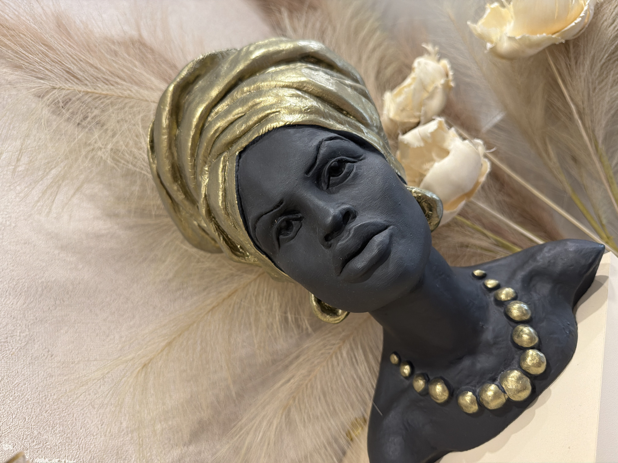 An eye-level shot captures a stylized bust of a person, likely a woman, featuring a dark, matte grey or black complexion. The figure wears a prominent gold headwrap, intricately sculpted with folds that gleam with a metallic finish. Gold hoop earrings adorn the ears, and a chunky, textured gold beaded necklace rests around the neck, contrasting sharply with the dark skin. The facial features are distinct, with a serious, contemplative expression.
The bust is positioned against a soft, light-colored background composed primarily of numerous feathery plumes, possibly pampas grass, creating a delicate and textured backdrop. Interspersed among the plumes are several cream or light yellow flowers, which appear to be dried or artificial, adding a subtle organic element to the scene. The overall color palette is elegant, dominated by the rich contrast of dark grey/black and metallic gold against the warm, neutral tones of the background. The lighting is soft and even, highlighting the textures of the sculpture and the surrounding decor. The image depicts a decorative display, likely in an indoor setting such as a home, gallery, or retail space in Dzerzhinsk, Russia. There is no discernible activity or visible text.