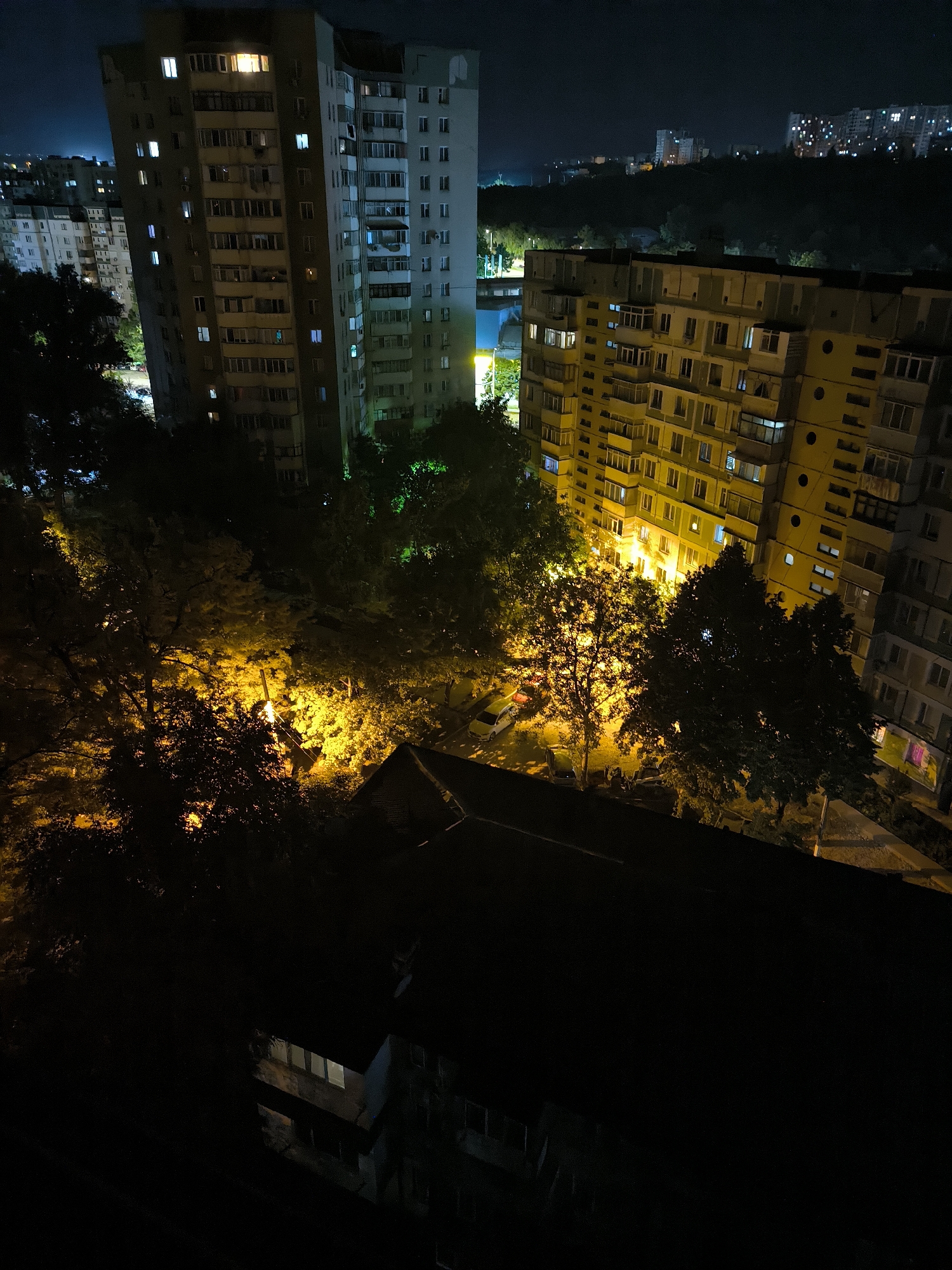 This image depicts a nighttime view of apartment buildings and trees in a city. The scene is illuminated by streetlights and the lights from the apartment windows, creating a contrast between the dark sky and the illuminated structures. The apartment buildings are of varying heights and architectural styles, with some appearing to be Soviet-era panel buildings. Trees with foliage are visible in the foreground and mid-ground, some of which are lit by yellow streetlights, casting an orange glow on the surroundings. In the background, other buildings and lights suggest a wider urban landscape. The image appears to be taken from an elevated perspective, looking down onto the scene. Based on the architectural style of the buildings and the overall urban setting, this scene is likely located in Chișinău, Moldova.