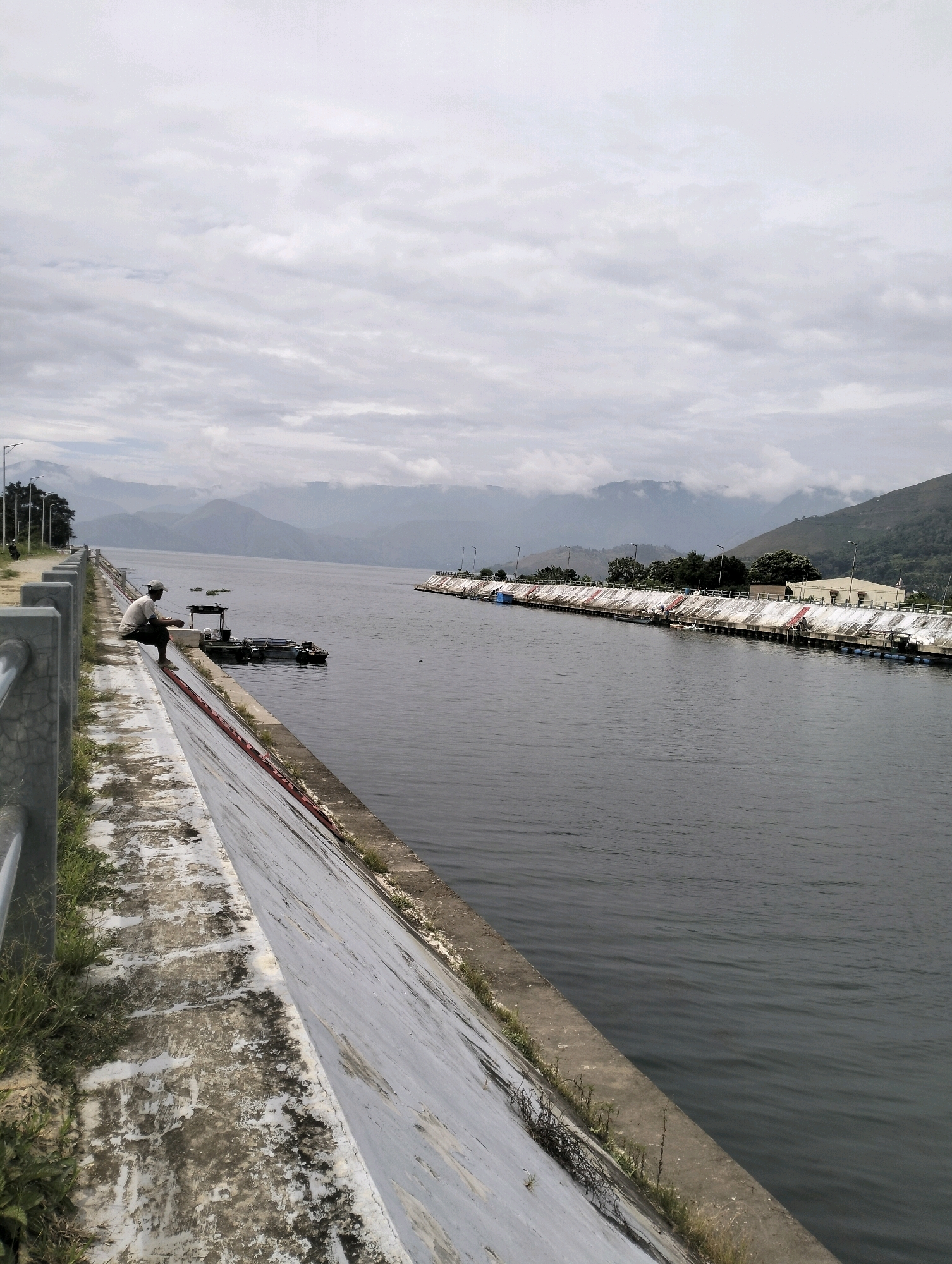 The image captures a view of a lake or river with a concrete embankment along its edge in Pangururan, Indonesia. On the left side of the embankment, a person is seated, facing away from the camera, looking out at the water. A railing runs along the top of the embankment. Several boats are visible on the water, some docked along the opposite bank, which features structures and vegetation. In the background, mountains are shrouded in mist under a cloudy sky. The overall scene depicts a serene, overcast day by the water.