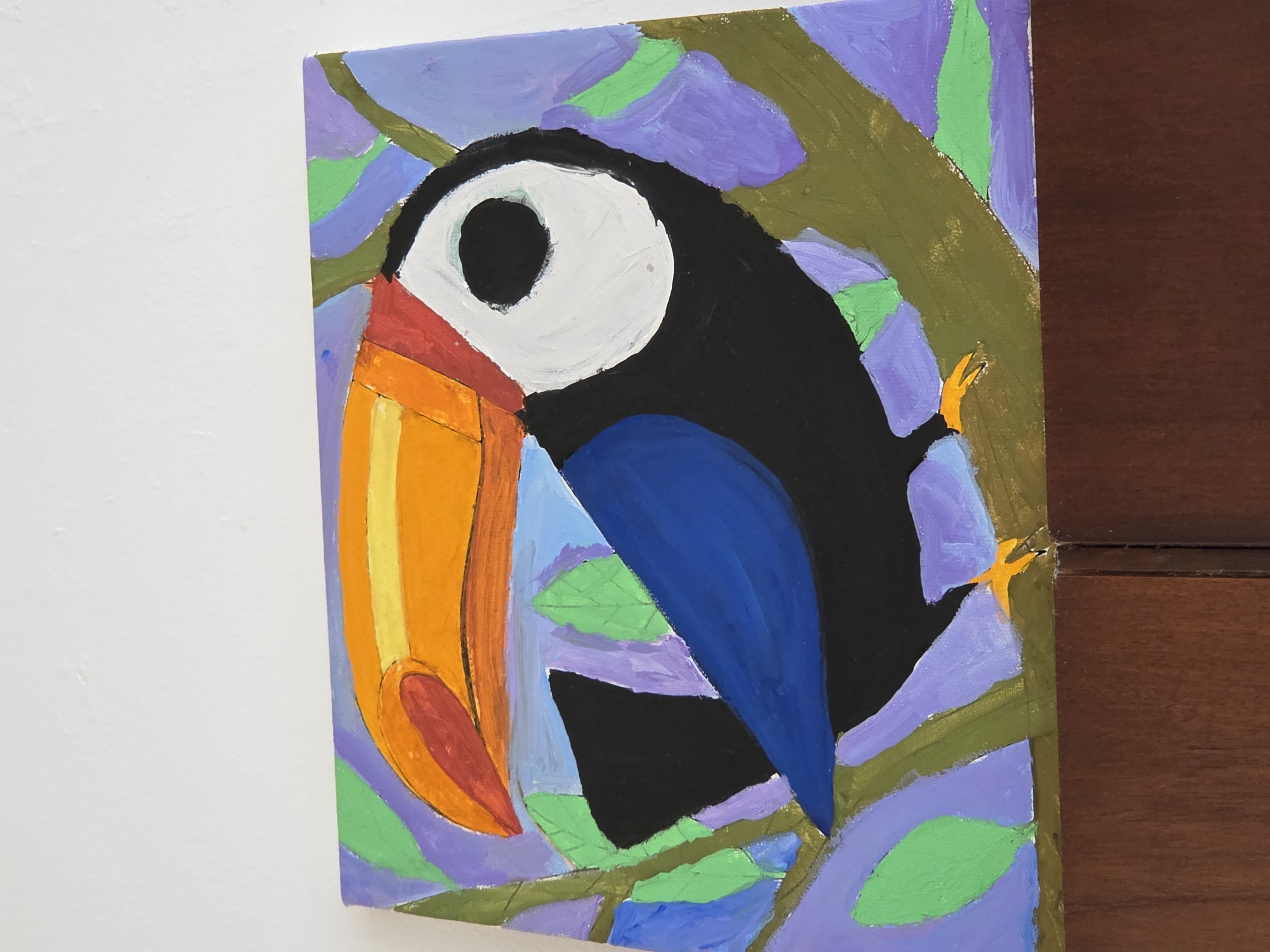 A close-up shot shows a painting of a toucan perched in a tree. The painting is in a naive, stylized style, with bold colors and simple shapes. The toucan has a large orange beak with a red tip, a black head and body, and a blue wing. The tree branches are olive green, and the leaves are a mix of purple, blue, and green. The painting is mounted on a wall, and part of a dark brown wooden panel is visible in the right of the frame. The background is light-colored, presumably another wall. The image does not provide any information to verify if the painting or the location is in North Kuta, Indonesia.