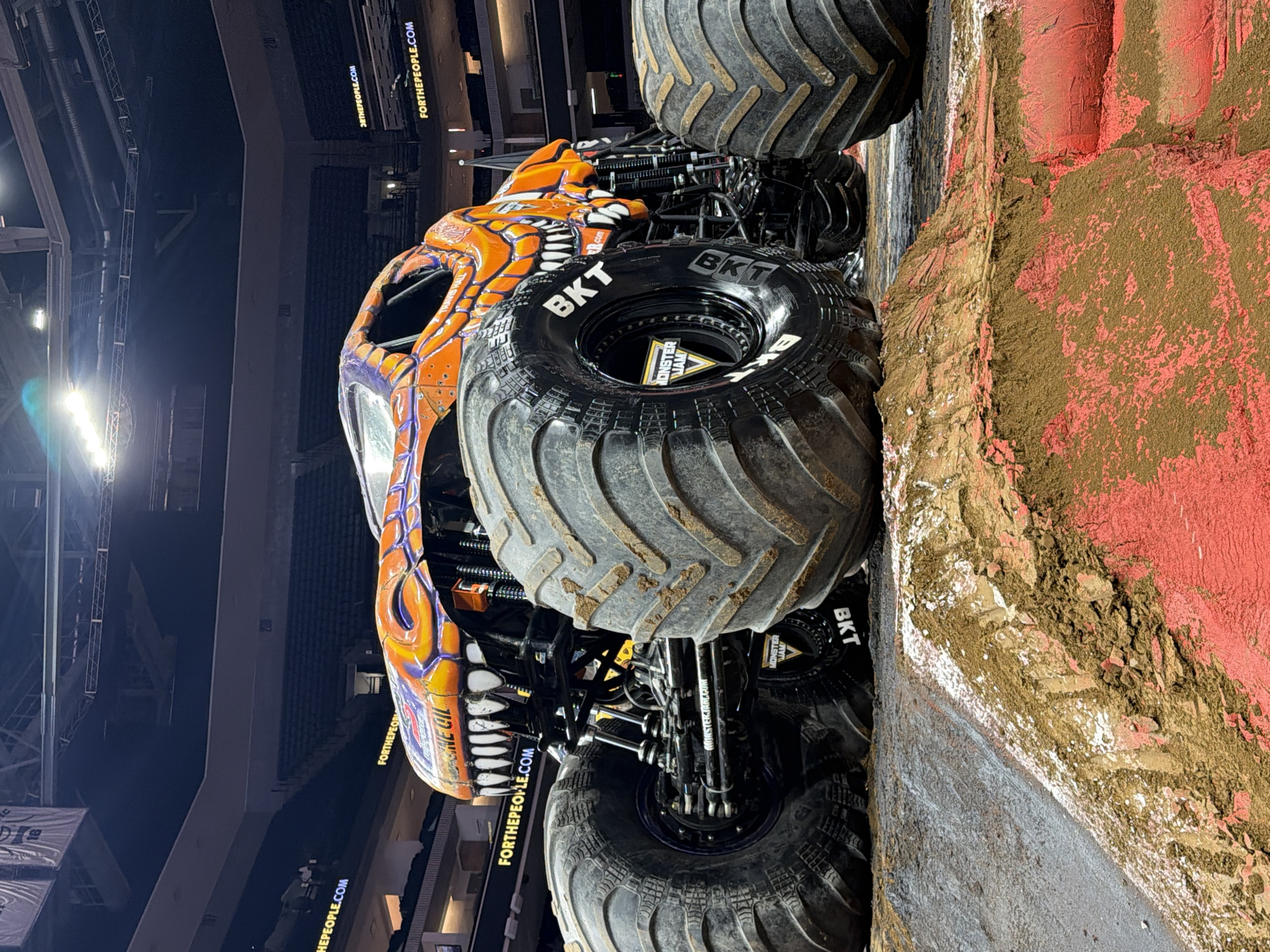 A monster truck is parked on a dirt ramp inside an arena. The truck is orange with purple accents and has a dinosaur-like design with sharp teeth. The words "FOR THE PEOPLE.COM" are visible on banners in the background. The arena appears to be the Golden 1 Center in Sacramento, California, United States of America. The lighting suggests it is nighttime and there are no people visible in the immediate vicinity of the truck, though some spectators are visible in the upper sections of the arena.