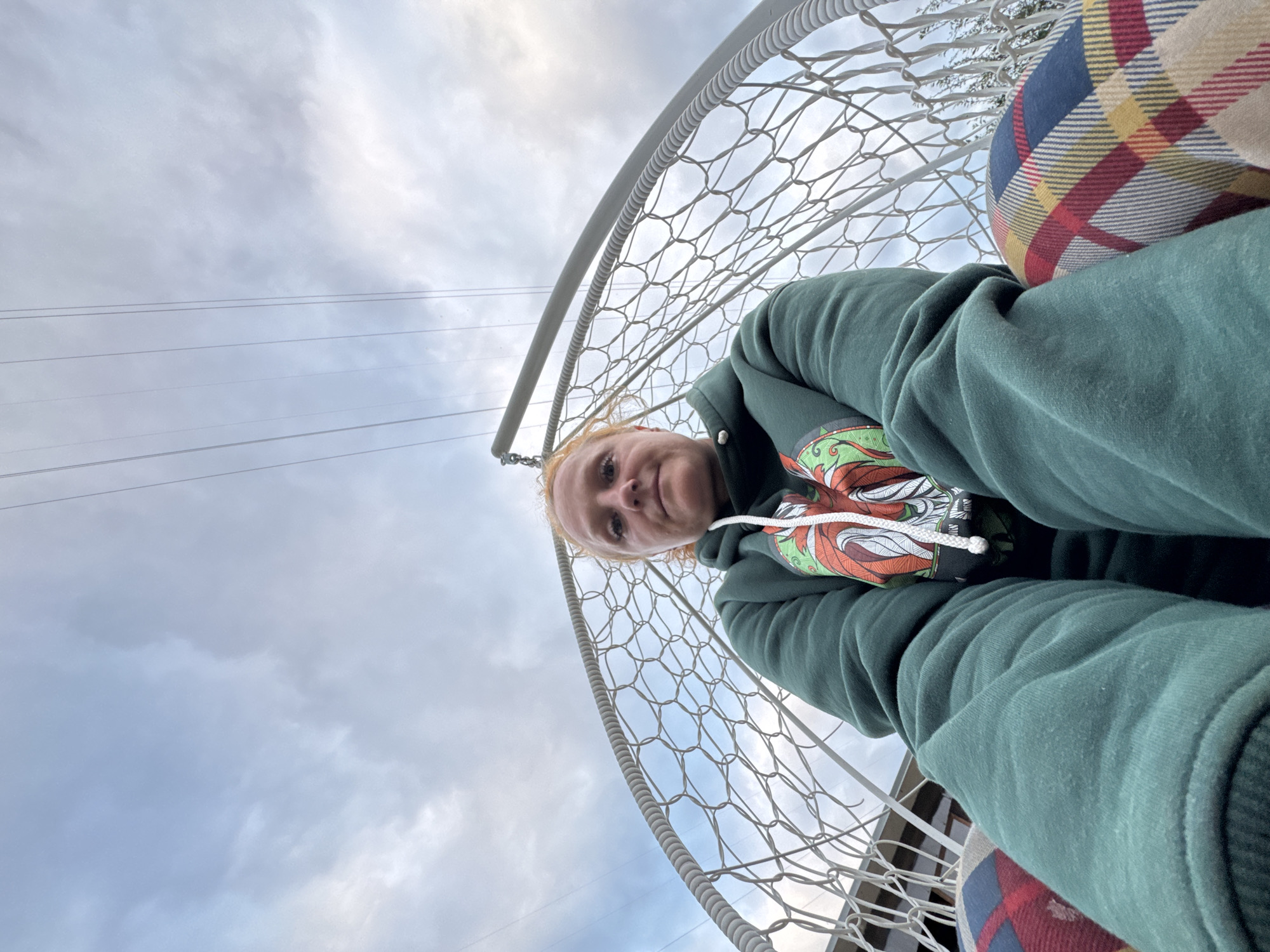 In an outdoor, low-angle, medium shot, a woman with red hair, wearing a dark green hooded sweatshirt with a graphic print and plaid leggings, is seated in a white hanging chair with a netted seat and back. She is looking towards the camera. The background consists of a cloudy sky with visible power lines crisscrossing overhead. The scene appears to be in Chaikovsky, Russia.