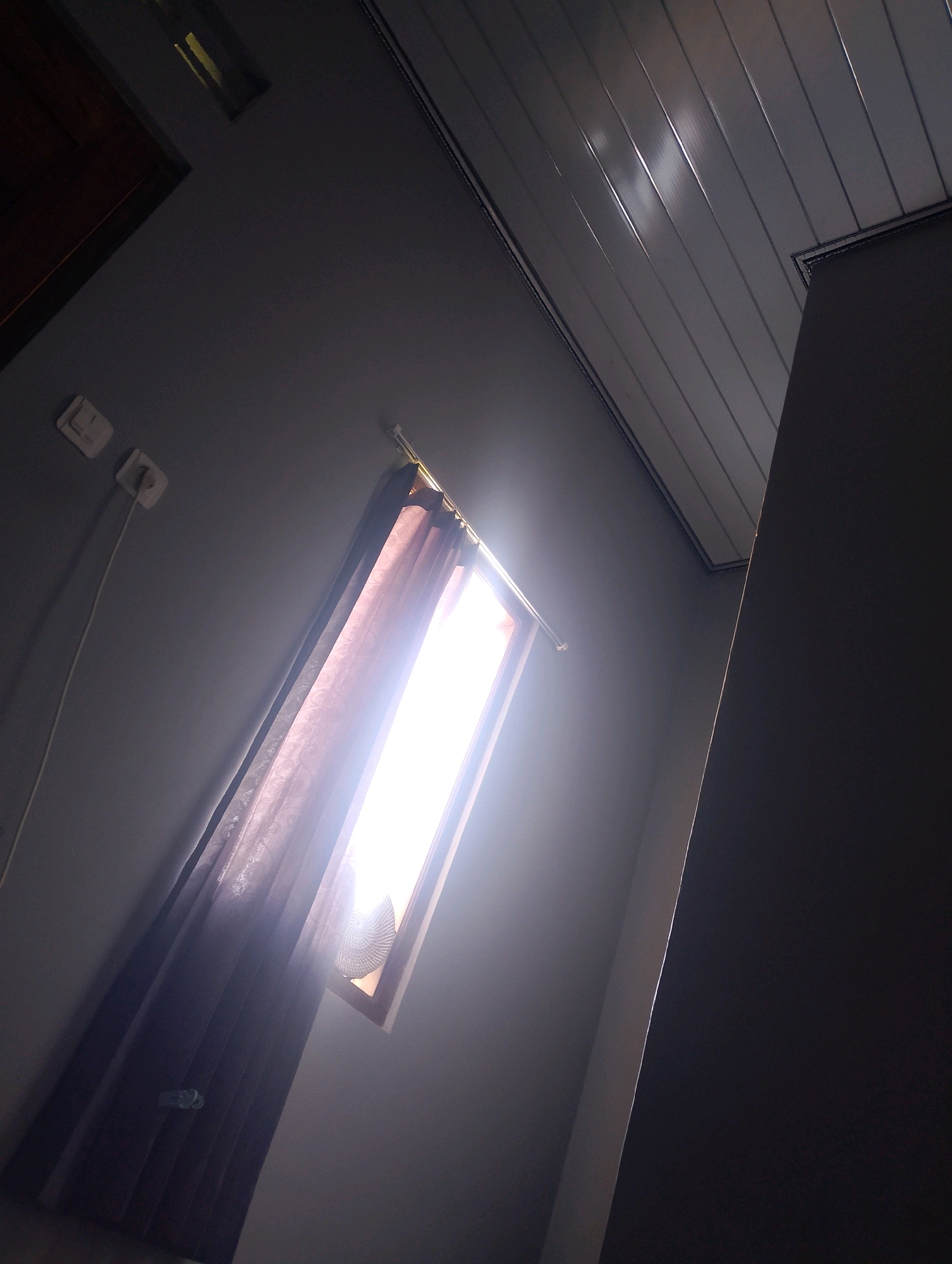 The image depicts an indoor scene with a strong light source coming from a window covered by translucent curtains. The light illuminates the curtains, creating a bright glow. The room appears to be a bedroom or living space, with a textured ceiling made of interlocking panels and a dark wall. On the wall, two electrical outlets are visible, one with a white cord plugged in. A portion of a dark, tall structure, possibly a wardrobe or cabinet, is visible on the right side of the frame, casting a shadow. The perspective suggests the photo was taken from a low angle, looking upwards. The scene is likely located within a residential property in Magelang, Indonesia, a city known for its diverse housing and architecture. The overall ambiance is one of natural light entering a dimly lit interior.