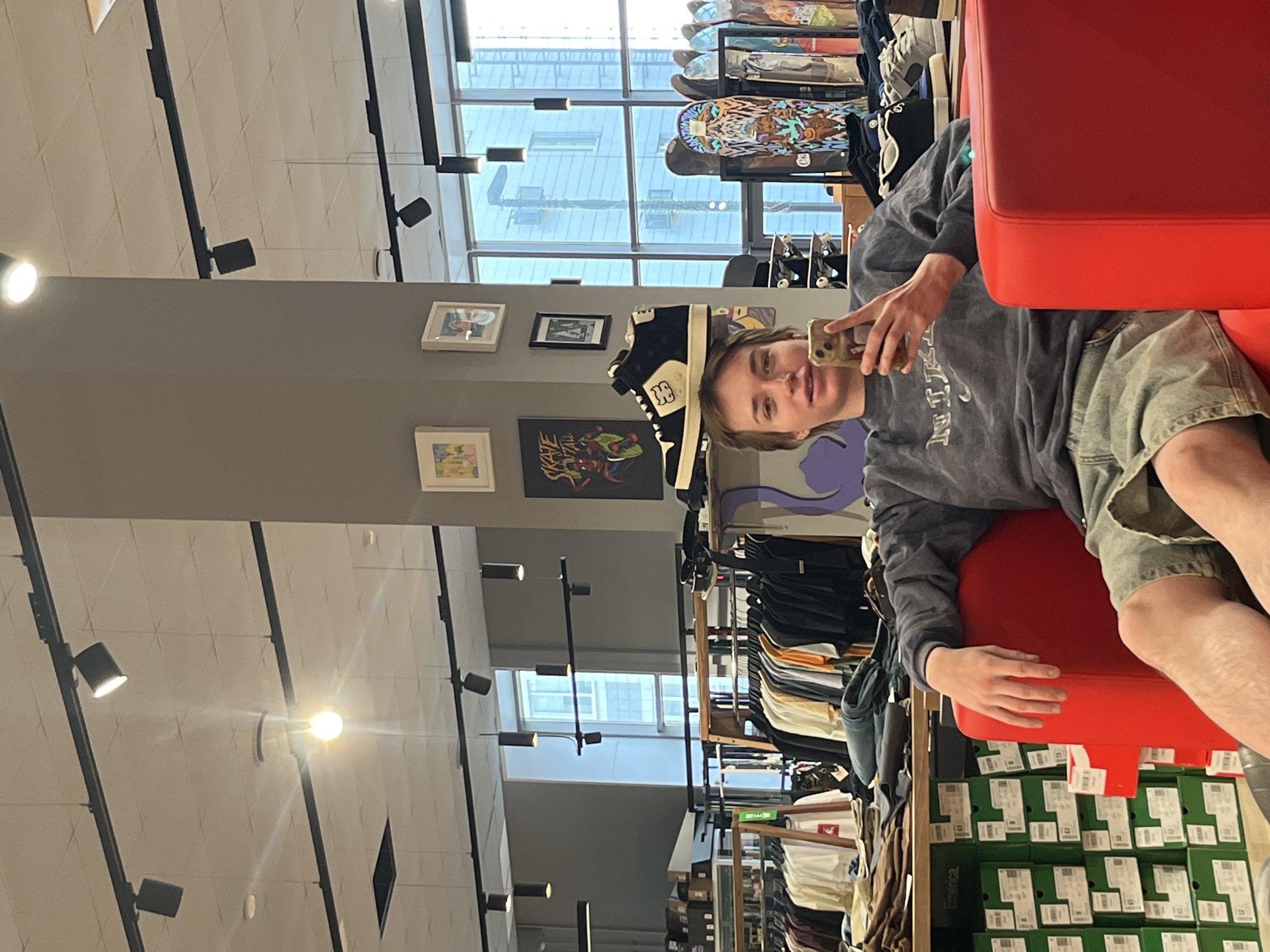 A person sits in a red chair inside a store. The individual is holding a phone and appears to be taking a selfie. The store is selling clothing, shoes, and skateboards. The location is likely Krasnoyarsk, Russia, based on the information available.