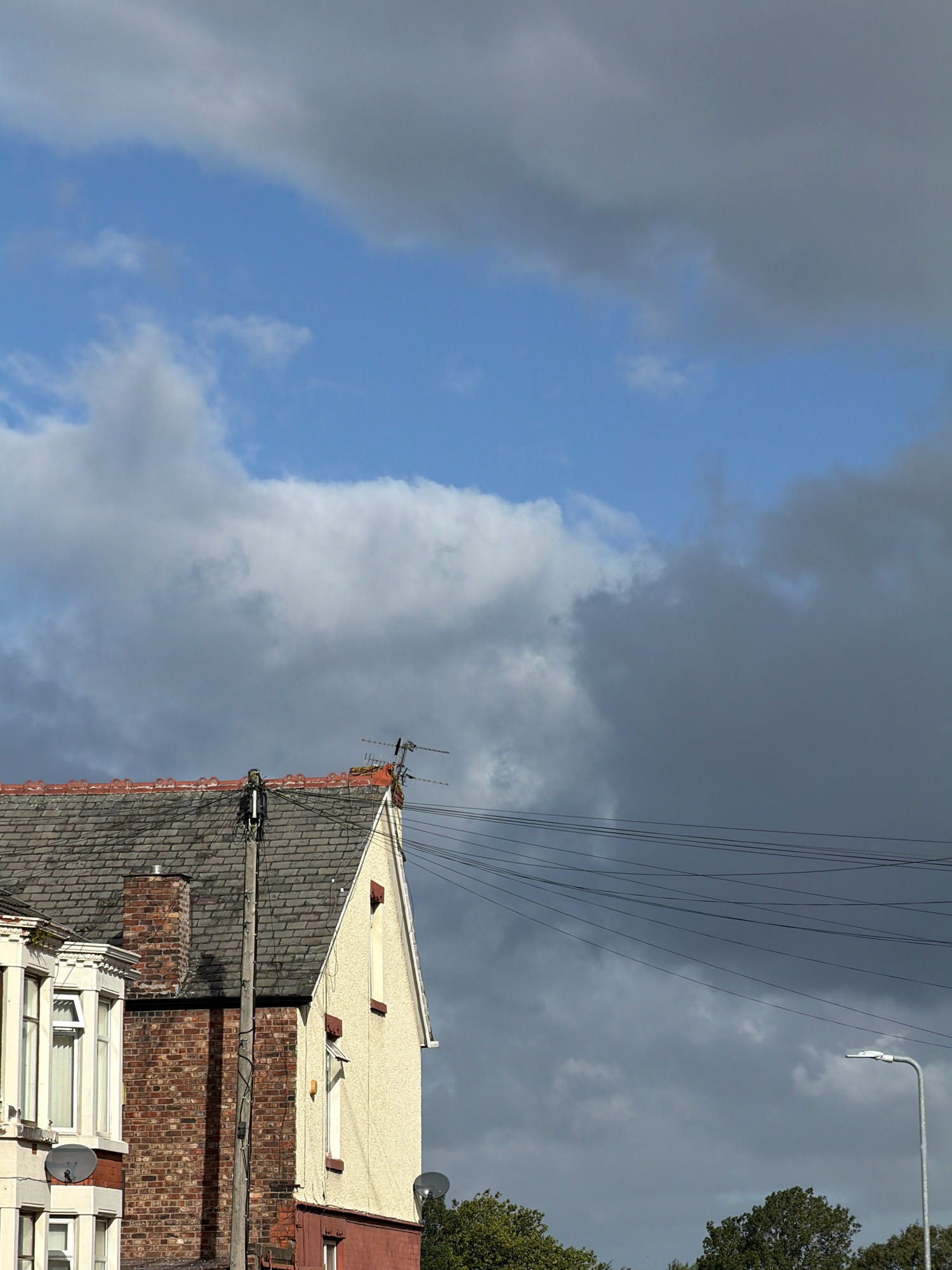 The image depicts a cloudy sky above a row of houses and a lamppost in an urban setting. The sky is a mix of blue and dark grey clouds. On the left, a portion of a house is visible with a bay window and brickwork. Next to it is a house with a cream-colored facade and a red base, featuring a prominent chimney and roof tiles. Electrical wires crisscross the scene, suspended between poles. A lamppost stands on the right, and trees are visible at the bottom. The overall impression is that of a typical residential street under a changeable sky.