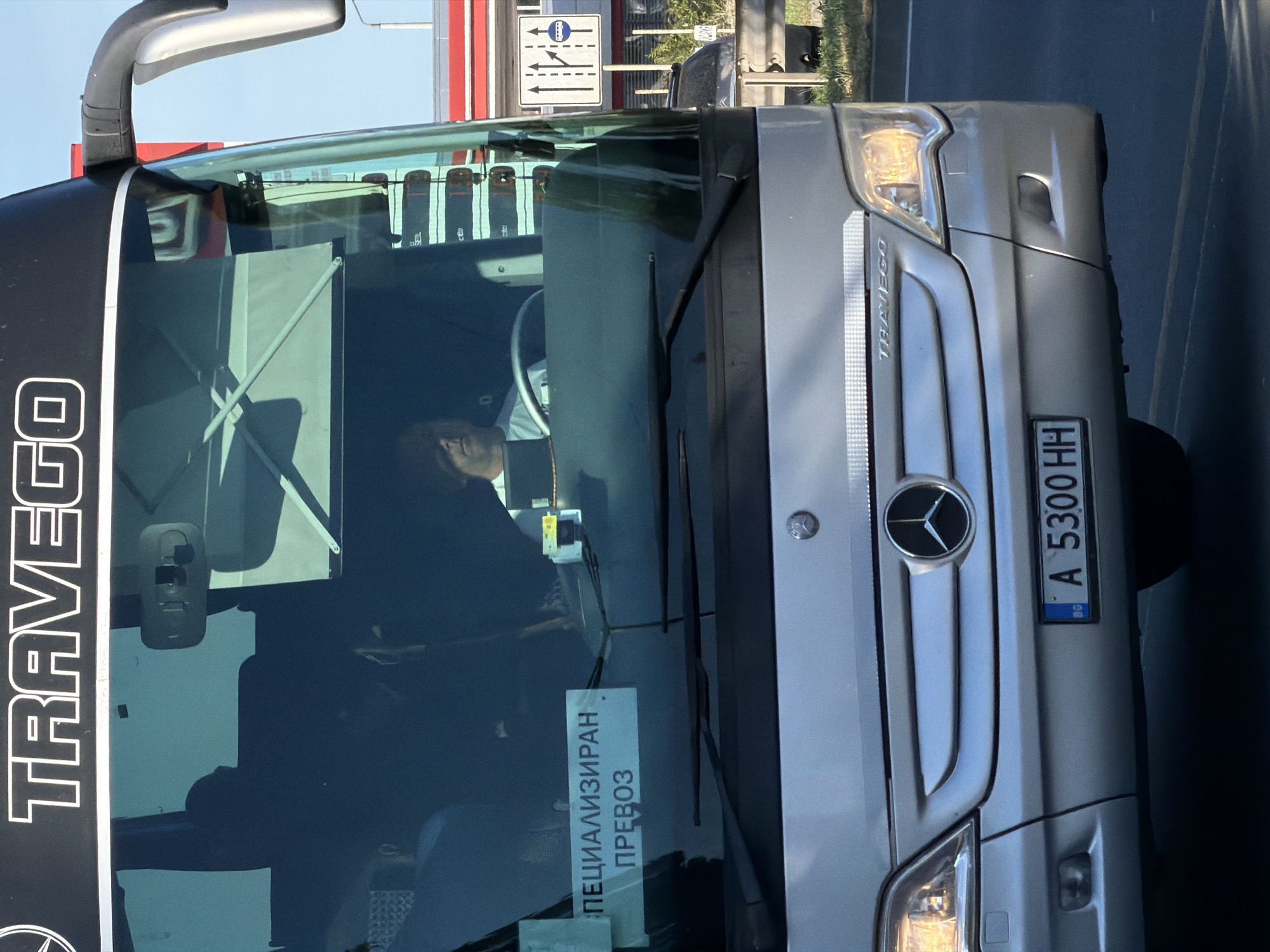 The image shows the front of a silver Mercedes-Benz Travego bus. The bus is parked on a street, likely during the daytime, given the bright sunlight. There is a sign with traffic directions visible in the background. On the windshield of the bus, a sticker with Cyrillic text is visible, reading "СПЕЦИАЛИЗИРАН ПРЕВОЗ", which translates to "SPECIALIZED TRANSPORT". A license plate is also visible on the front of the bus, displaying "A 5300 HH" and a blue European Union band. The driver of the bus is partially visible through the windshield, appearing to be a man. The overall scene suggests a bus ready for or having completed a transport service.