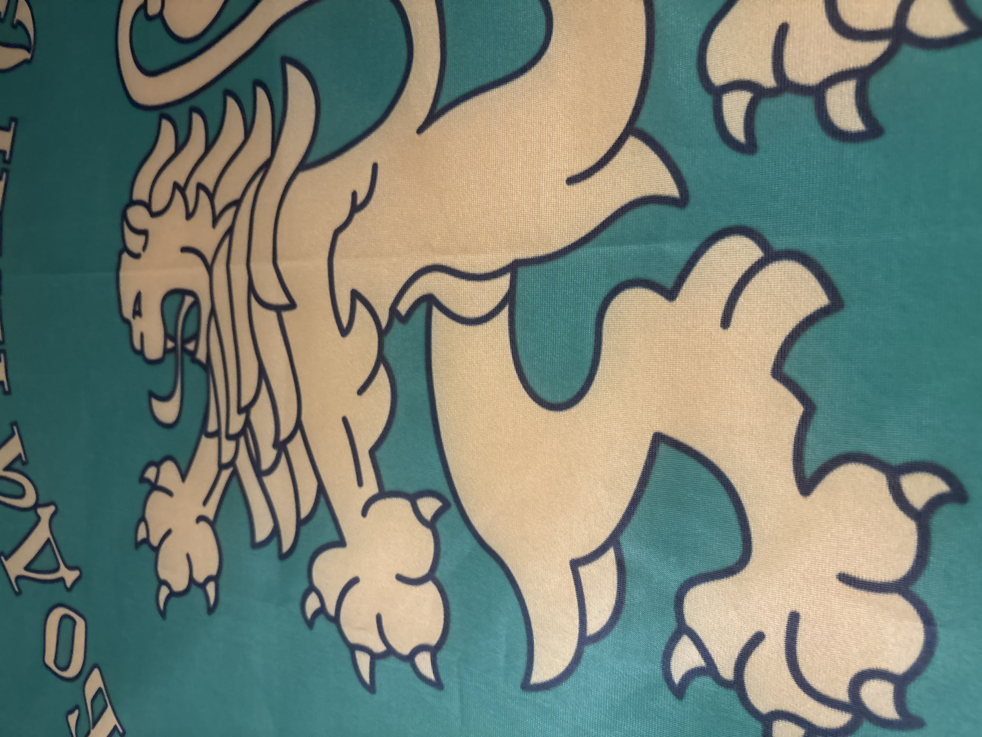 The image displays a close-up, angled shot of a flag featuring a stylized golden lion with a dark outline against a teal background. The lion is depicted in profile, with its mouth open and mane flowing. Partially visible at the top of the image are Cyrillic letters in white with a dark outline. This flag is identified as the flag of Plovdiv, Bulgaria. The specific location within Plovdiv is not discernible from the image itself. The material of the flag appears to be fabric with a visible weave.