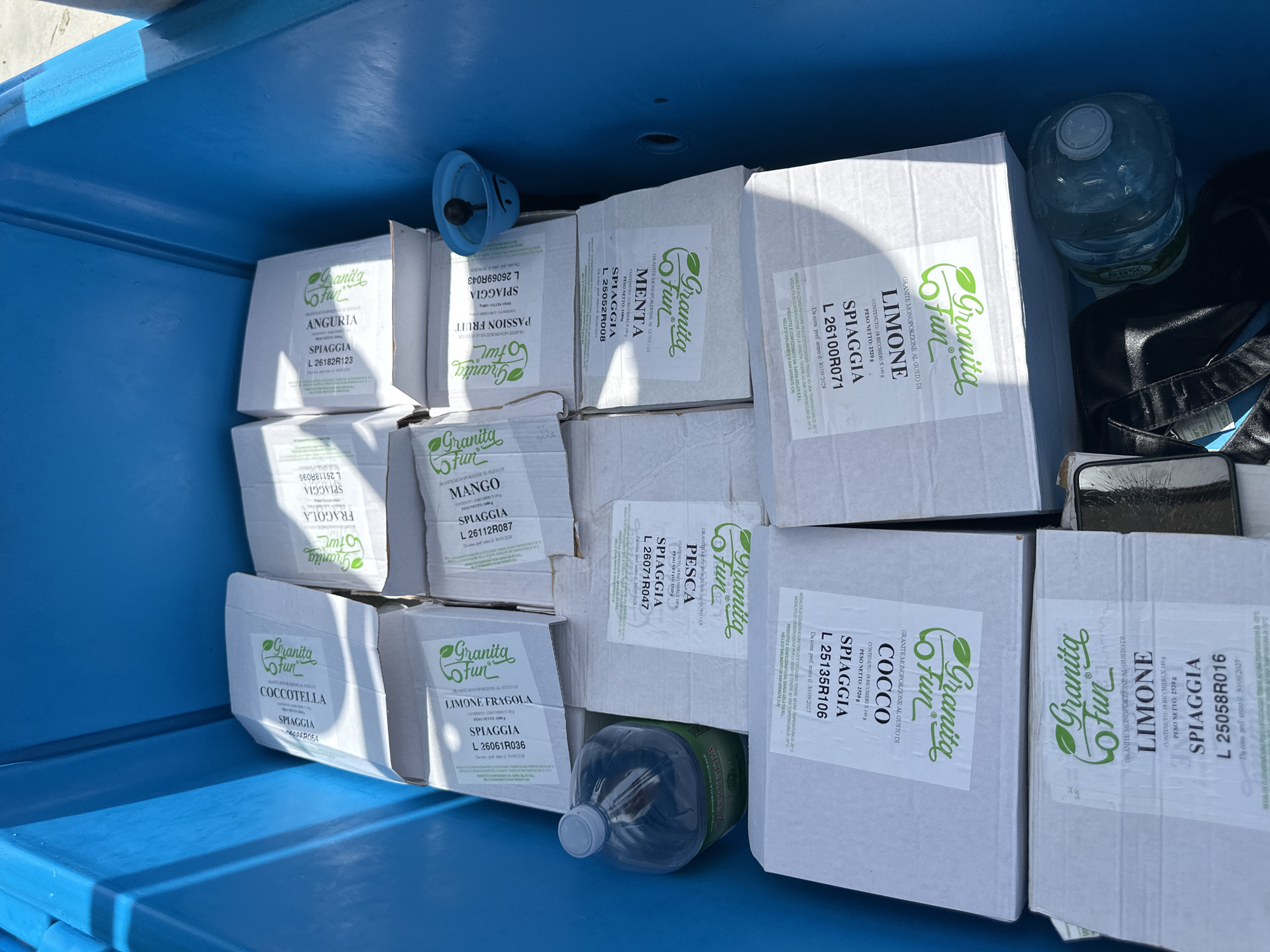 The image shows a close-up, high-angle view of the inside of a blue plastic container, likely a cooler or storage bin. The container is filled with numerous cardboard boxes, most of which are white with green and black text and graphics. The boxes appear to contain individually packaged items, possibly food or beverages, with labels indicating flavors such as "Fragola" (Strawberry), "Anguria" (Watermelon), "Limone" (Lemon), "Menta" (Mint), "Cocco" (Coconut), and "Pesca" (Peach). Some boxes also have text like "Granita Fun" and "SPAGGIÀ".
In the lower left corner, a large plastic bottle of water with a blue cap is visible. To the right of the water bottle and slightly behind it, a dark-colored bag or backpack is partially visible. In the upper right quadrant, a small blue plastic object with a smiley face design is resting on the edge of the container. The lighting suggests an outdoor, daytime setting, likely in a sunny environment, as indicated by the bright illumination and shadows. The blue container itself appears to be in good condition, with some scuff marks visible on its inner surface. The overall scene depicts a collection of packaged goods stored within a portable container, suggesting preparation for an event or transport. The location is identified as Crabonaxa/Villasimius, Italy, which provides a geographical context to the scene.