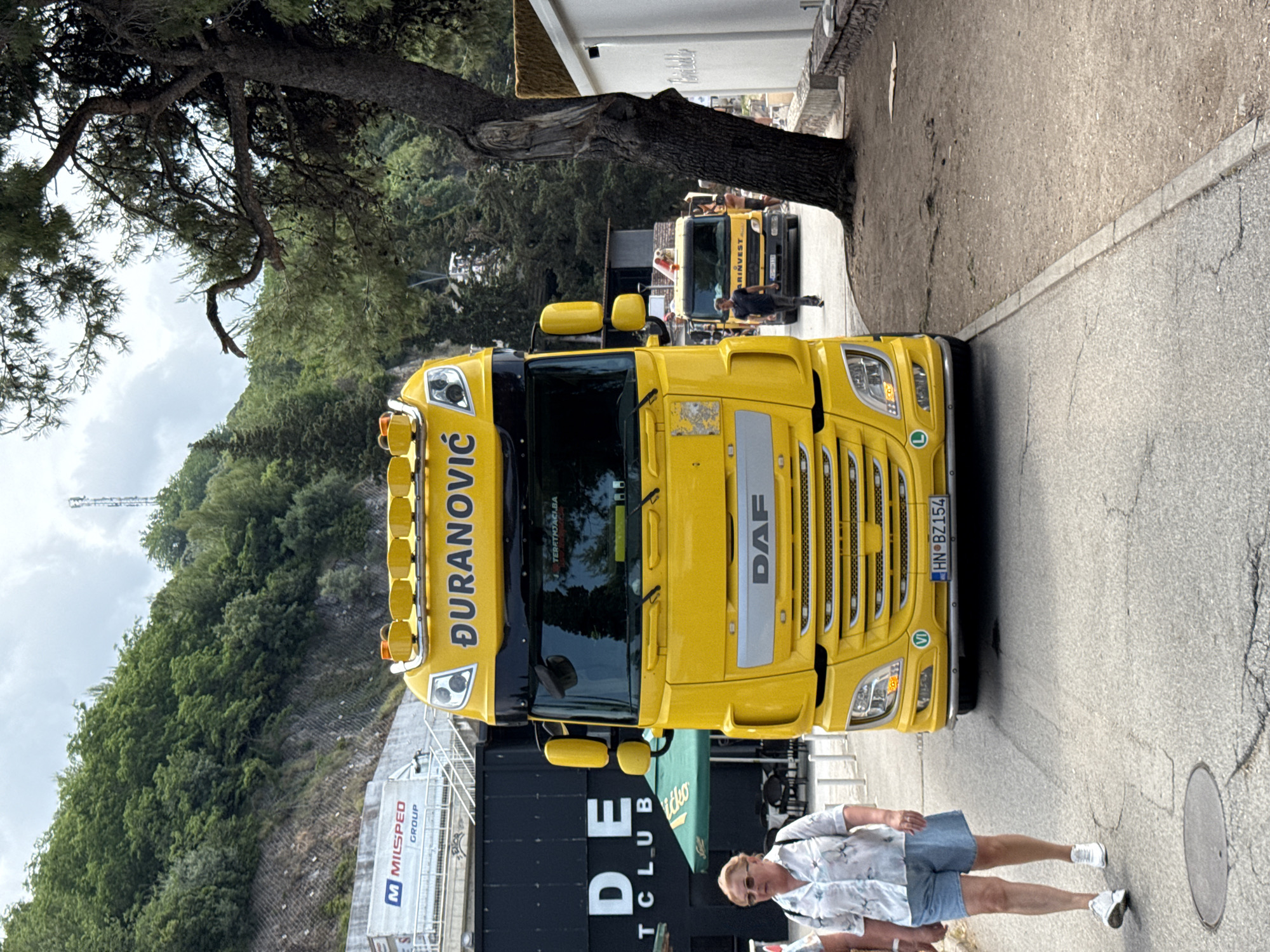 This image, set in Budva, Montenegro, captures a daytime scene on a sloped asphalt road amidst lush green surroundings. The sky appears overcast with visible clouds, suggesting moderate lighting.
In the foreground, a bright yellow DAF semi-truck cab is prominently featured, facing directly towards the viewer. The name "ĐURANOVIĆ" is displayed across the top of its cab, and "DAF" is visible on its front grille. Its license plate, "HN BZ154," indicates registration in Herceg Novi, Montenegro. On the truck's windshield, the text "TERETNJACI.BA" is visible. The truck also has additional lights mounted on its roof.
To the right of the large truck, a man is walking on the road. He wears a light-colored, patterned short-sleeved shirt, denim shorts, and white sneakers, with sunglasses resting on his head. In the background, further down the road, a smaller yellow truck with the word "HARINVEST" on its side can be seen, with another man standing near it.
The setting is characterized by a mix of natural and built elements. A large tree with a thick, textured trunk and extensive branches dominates the upper left side of the frame. A dense, verdant hillside covered in various trees and foliage rises in the background. On the left side, behind the large truck, parts of structures are visible, including signage that reads "M MILSPED GROUP" and "DE CLUB," with a green canopy partially displaying the word "JATKO." On the right, a white building with a distinctive thatched roof element is partially visible. The asphalt road exhibits some cracks, and a circular manhole cover is noticeable near the walking man. The scene depicts a slice of daily activity, likely related to commerce or logistics, integrated into the natural landscape of the region.