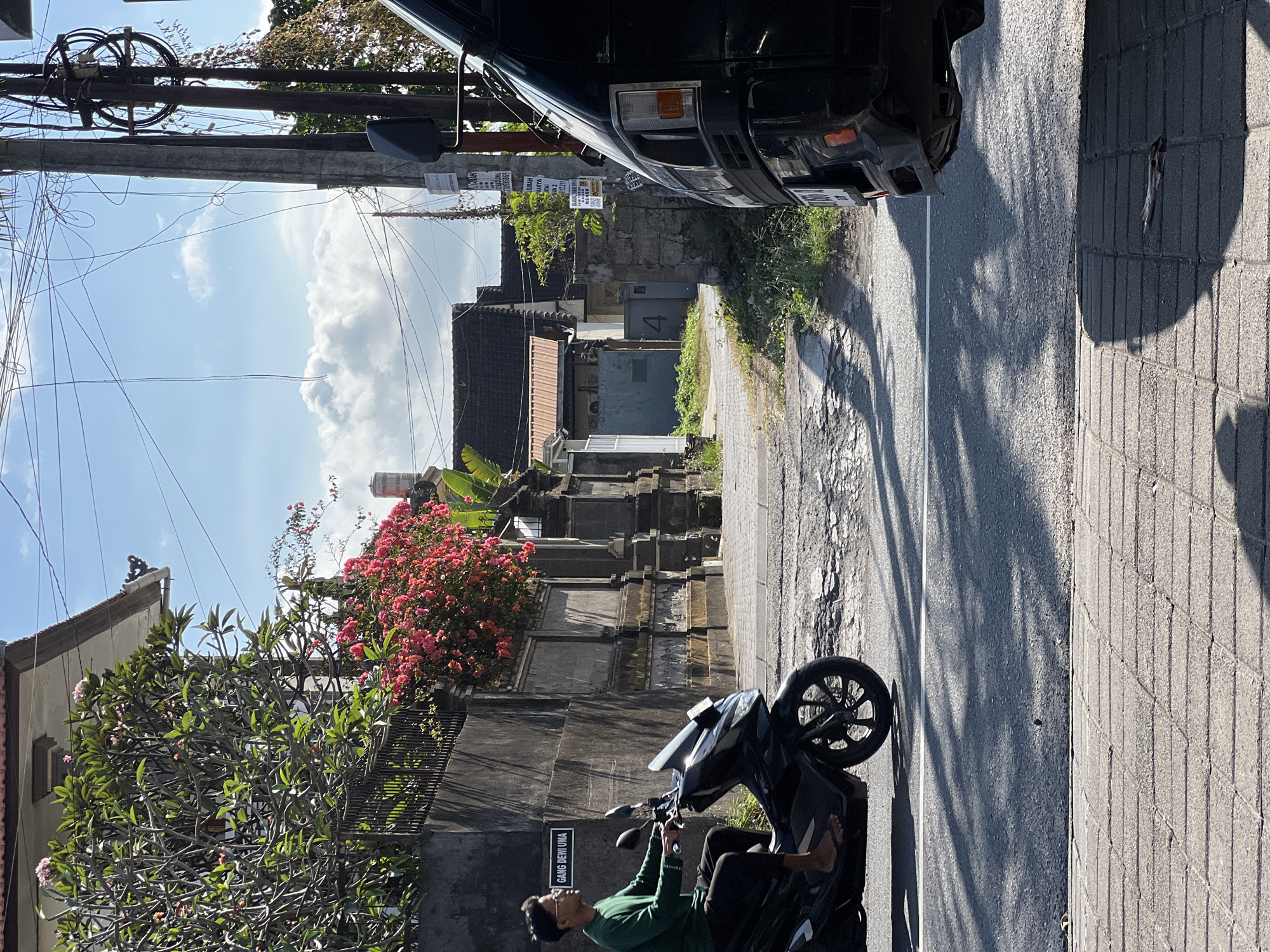 In Denpasar, Indonesia, a young man wearing glasses and a green long-sleeved shirt rides a black scooter on a street lined with traditional Balinese architecture. The scooter's front wheel is on the left side of the frame, while the man's legs are visible, wearing dark pants and flip-flops. To the right, the back of a dark-colored van is parked, partially obscuring the view of the street and the man. The street itself is paved with asphalt and shows signs of wear, with patches of damaged surface.
The environment features a high stone wall with an ornate railing on the left, adorned with vibrant pink bougainvillea flowers. A sign on the wall reads "GANG DEWI UMA," indicating the alley's name. Behind the wall, parts of houses with terracotta roofs are visible. Tall utility poles with a tangle of electrical wires stretch across the bright blue sky, which is dotted with fluffy white clouds. The sunlight casts long shadows on the street and the tiled sidewalk in the foreground, suggesting it is late afternoon. The scene captures a typical street view in a residential area of Denpasar, characterized by the combination of local architecture, vehicles, and the tropical flora.