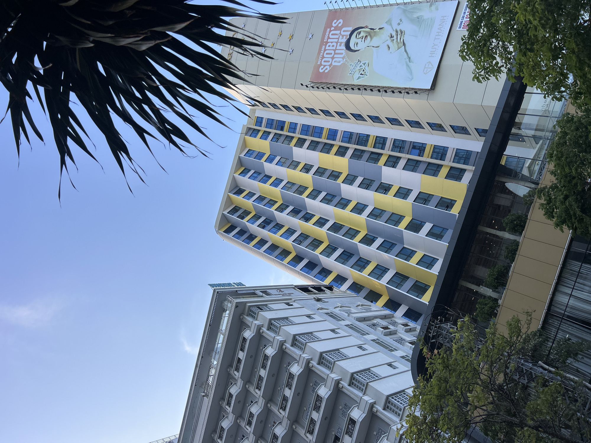 The image shows a low-angle view of several buildings in Nha Trang, Vietnam. The sky is clear and blue. A palm tree's fronds are visible in the upper left corner. The tallest building has a billboard on top featuring a man's image and the text "SOOBIN'S QUEEN". The building is decorated with alternating yellow and grey panels around the windows. A second building is a light grey color and has ornate window treatments. Trees partially obstruct the view of the buildings on the right side of the frame.
