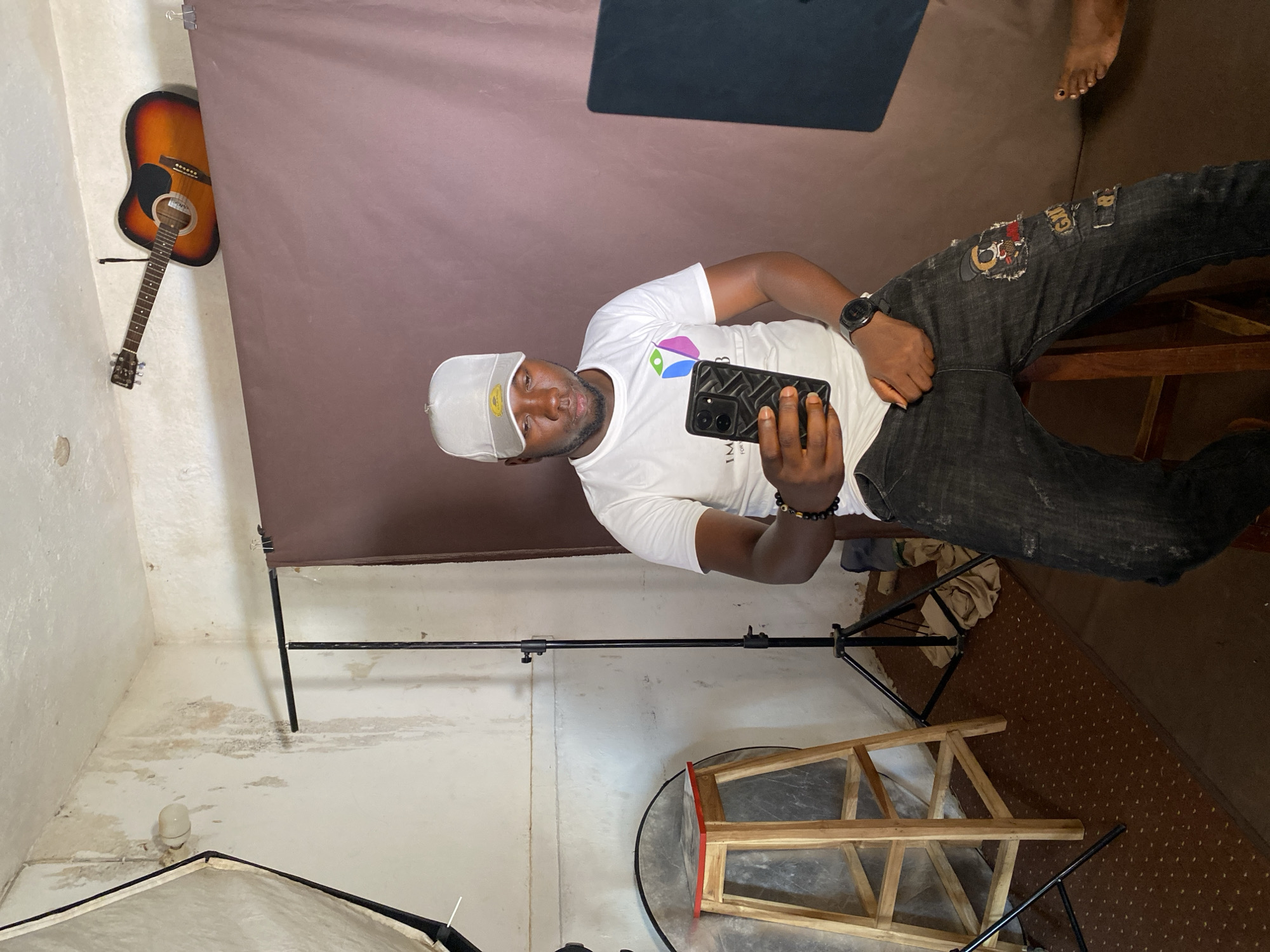 An image, rotated 90 degrees clockwise, shows a dark-skinned man in a casual indoor setting, likely a photography studio or makeshift photo area. The man, positioned centrally, is viewed from the chest up, with his full body visible from the waist down. He wears a white t-shirt with a colorful, abstract logo and partially visible text "IM" (likely part of "IMAGINATION"), dark ripped jeans with patches, a white baseball cap featuring a yellow smiley face emoji, and a black wristwatch on his left wrist. He holds a black smartphone with a textured case in his right hand, seemingly taking a selfie or looking at the screen. His expression is neutral.
Behind the man is a solid, dark reddish-brown backdrop, likely fabric, attached to a metal stand. To the left, a light-colored wall features an acoustic guitar hanging on it. The floor beneath the man transitions from a lighter, possibly concrete or tiled, area on the left (showing some water stains or discoloration) to a dark brown patterned carpet on the right. Various photography equipment, including metal stands and a partially visible white softbox or light diffuser, are scattered around the floor. A wooden frame, resembling a small stool or prop, is also visible. In the upper right corner, a bare foot, possibly belonging to another person or the subject's own foot seen at an odd angle due to the rotation, is visible on the carpeted floor. The overall impression is that of a prepared but somewhat unpolished photo shoot environment.