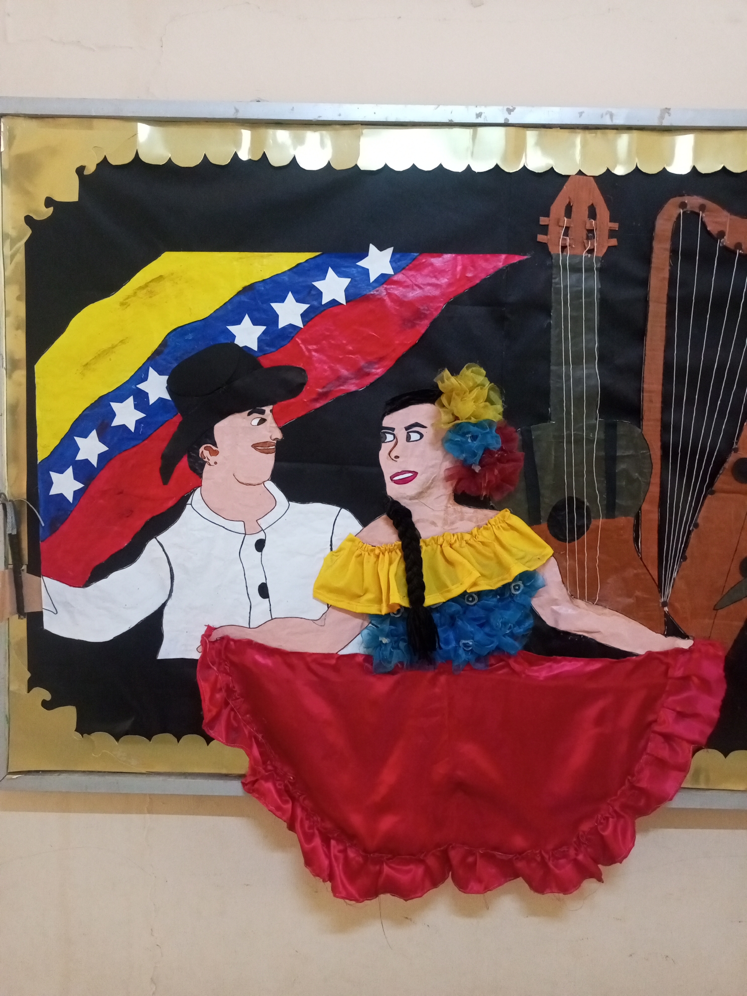 The image depicts a vibrant bulletin board display featuring a folk art representation of a man and a woman in traditional attire, set against a backdrop of the Venezuelan flag and musical instruments. The man, on the left, wears a black hat and a white shirt with black buttons. The woman, on the right, is adorned in a yellow off-the-shoulder top with blue frills and a large red skirt that drapes over the bottom of the display. Both figures have distinct facial features and appear to be interacting. Behind them, the Venezuelan flag is prominently displayed with its yellow, blue, and red stripes and white stars. To the right of the figures, parts of a guitar and a harp are visible, suggesting a cultural or musical theme. The display is framed by a golden scalloped border and a silver metal edge. The bulletin board itself is mounted on a beige wall, which shows signs of wear and discoloration, possibly in a classroom or public space. The overall impression is one of a festive cultural celebration, likely created as a decorative or educational exhibit.
The location is the bulletin board at the *Museo de Arte de Occidente* in Santa Ana, Venezuela.