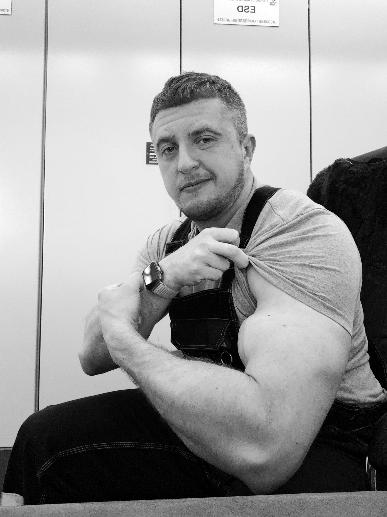 A black and white, medium shot shows a man sitting on a chair in an office-like location. He is wearing a grey t-shirt and dark-colored overalls. He is flexing his right arm and lifting the sleeve of his shirt to show his bicep. He is wearing a wristwatch on his left wrist. The background consists of a white wall with two signs on it, one visible and one partially visible. The upper sign reads "E2D" in reverse. The bottom sign's text is not clear. A furry object that may be a seat cover is behind the man on the right. He has short hair and a slight beard and mustache. The location is Unknown, Unknown.