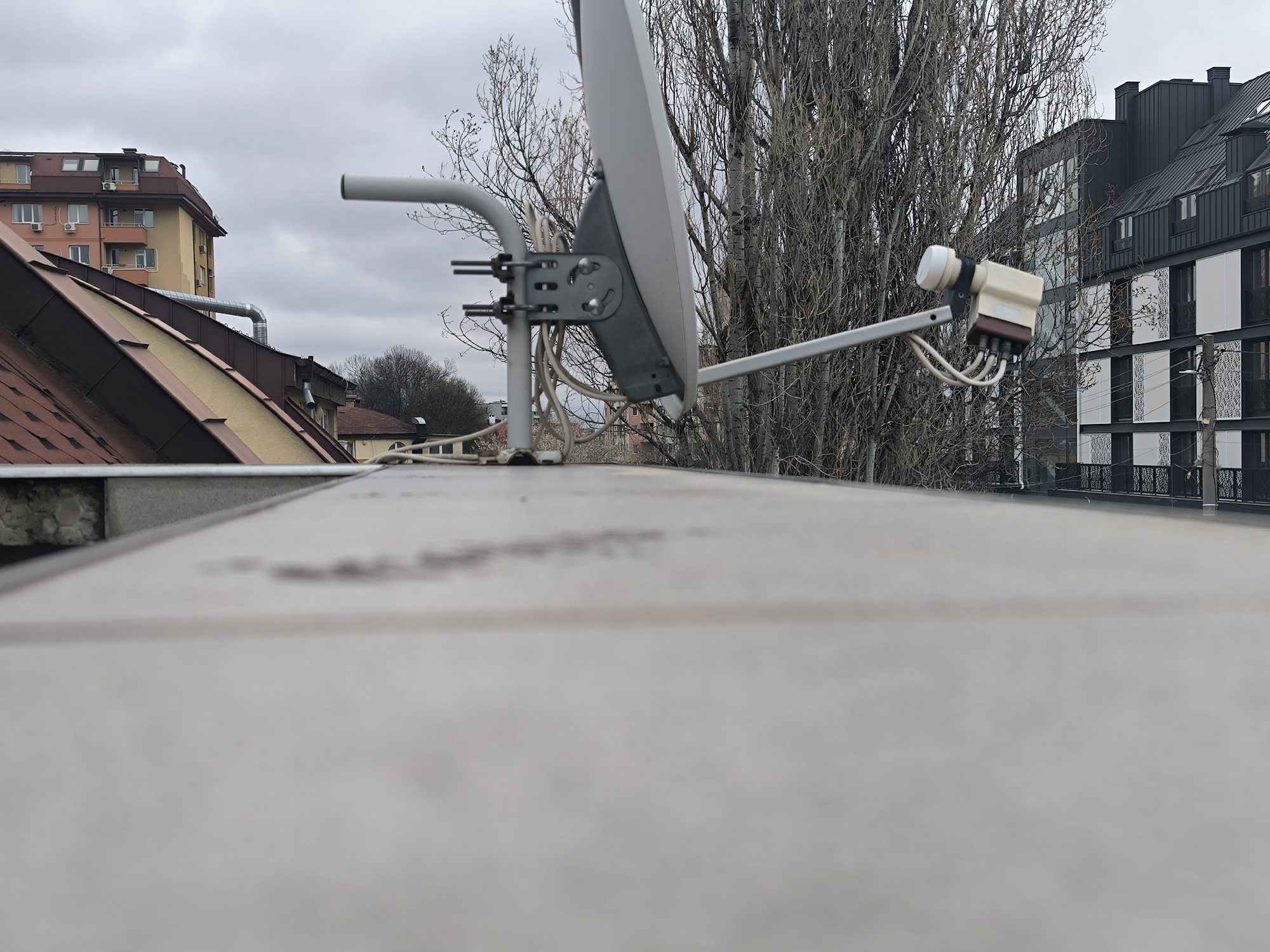 A satellite dish is mounted on a flat, tiled rooftop in Sofia, Bulgaria. The dish is grey with white wiring and appears to be aimed toward a modern building with black siding and white accents. Behind the dish, bare trees stand stark against an overcast sky. Other buildings are visible in the background, including one with orange and yellow facades and a brown tiled roof. The foreground is dominated by the close-up view of the rooftop surface.