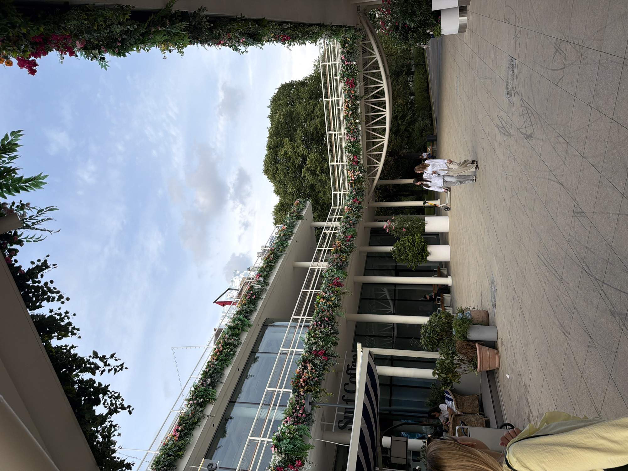 The image depicts the exterior of a contemporary building adorned with lush floral arrangements on its balconies and walkways, suggesting a location with a focus on aesthetics and outdoor space. The building features a modern architectural style with large glass windows and white railings. The surroundings include paved ground, greenery, and trees, hinting at a park-like or riverside setting. Two young women, dressed in casual white shirts and light-colored pants, are walking on the paved area. Another person is seated at a table outside what appears to be a cafe. The sky above is partly cloudy, indicating it is daytime. This scene is located at Gorky Park in Moscow, Russia.