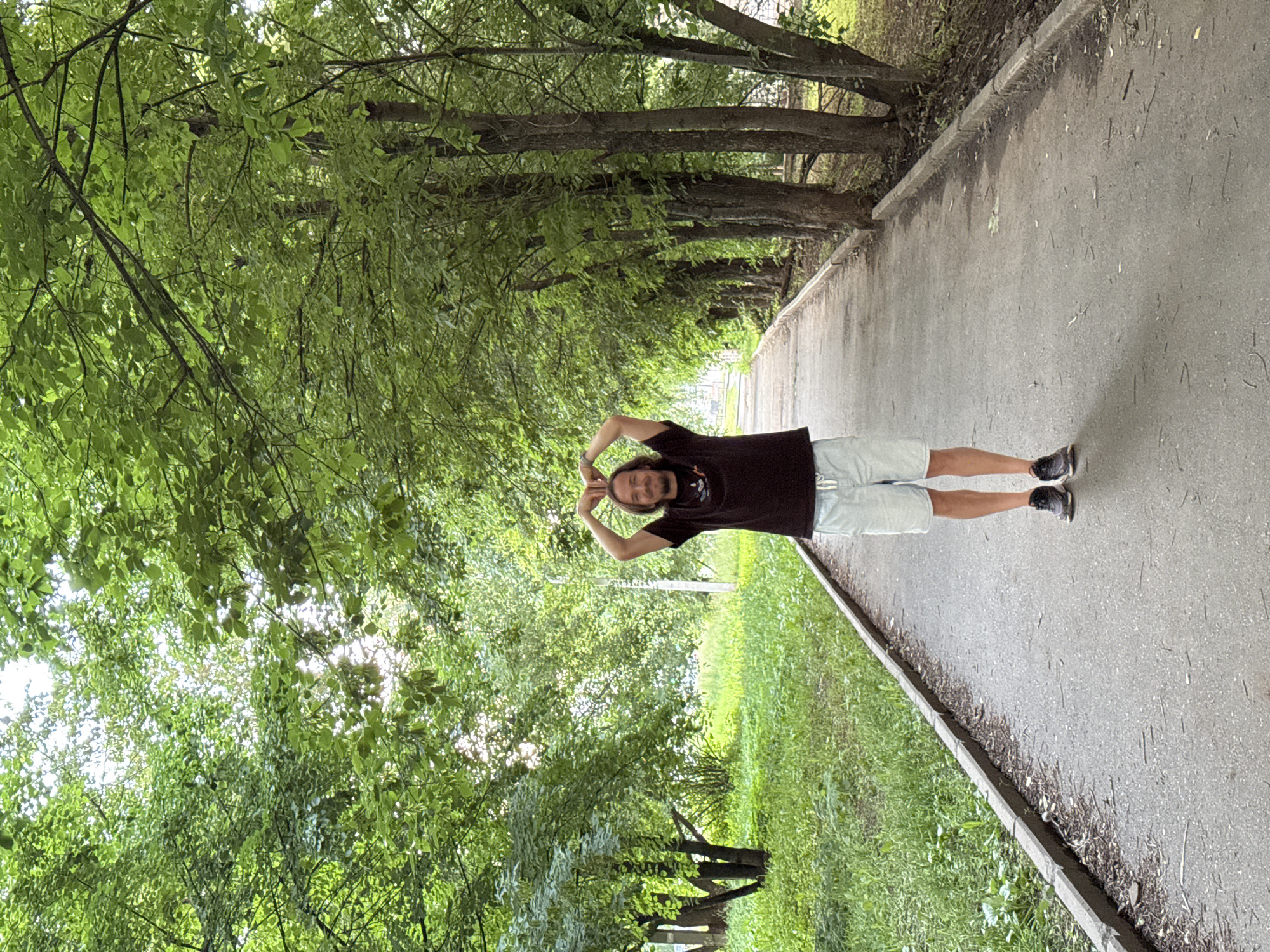 The image depicts a man standing on a paved path in a park-like setting. He is wearing a dark t-shirt and light-colored shorts, with athletic shoes on his feet. His hands are raised above his head, forming a heart shape. The path is lined on one side by a grassy area with trees, and on the other side by a row of trees. The trees are deciduous and their leaves are a vibrant green, suggesting a springtime or summer setting. Sunlight filters through the leaves, creating dappled light on the path and surrounding vegetation. The overall impression is of a peaceful, outdoor environment. While the image does not contain explicit markers, the context provided by the user suggests the location is the Izhevsk State Medical Academy park, located in the city of Izhevsk, Russia.