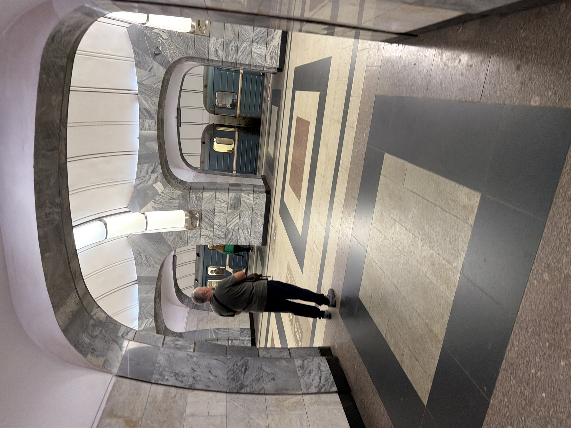 A man in a dark t-shirt and dark pants stands in a subway station, facing away from the camera. He has a bag slung over his left shoulder. The station features marble pillars and arched doorways. In the background, two subway cars are stopped at a platform, with a woman in a green jacket visible near the second car. The floor of the station is tiled with a pattern of light and dark squares and rectangles. The lighting appears to be artificial, emanating from long, overhead fixtures.