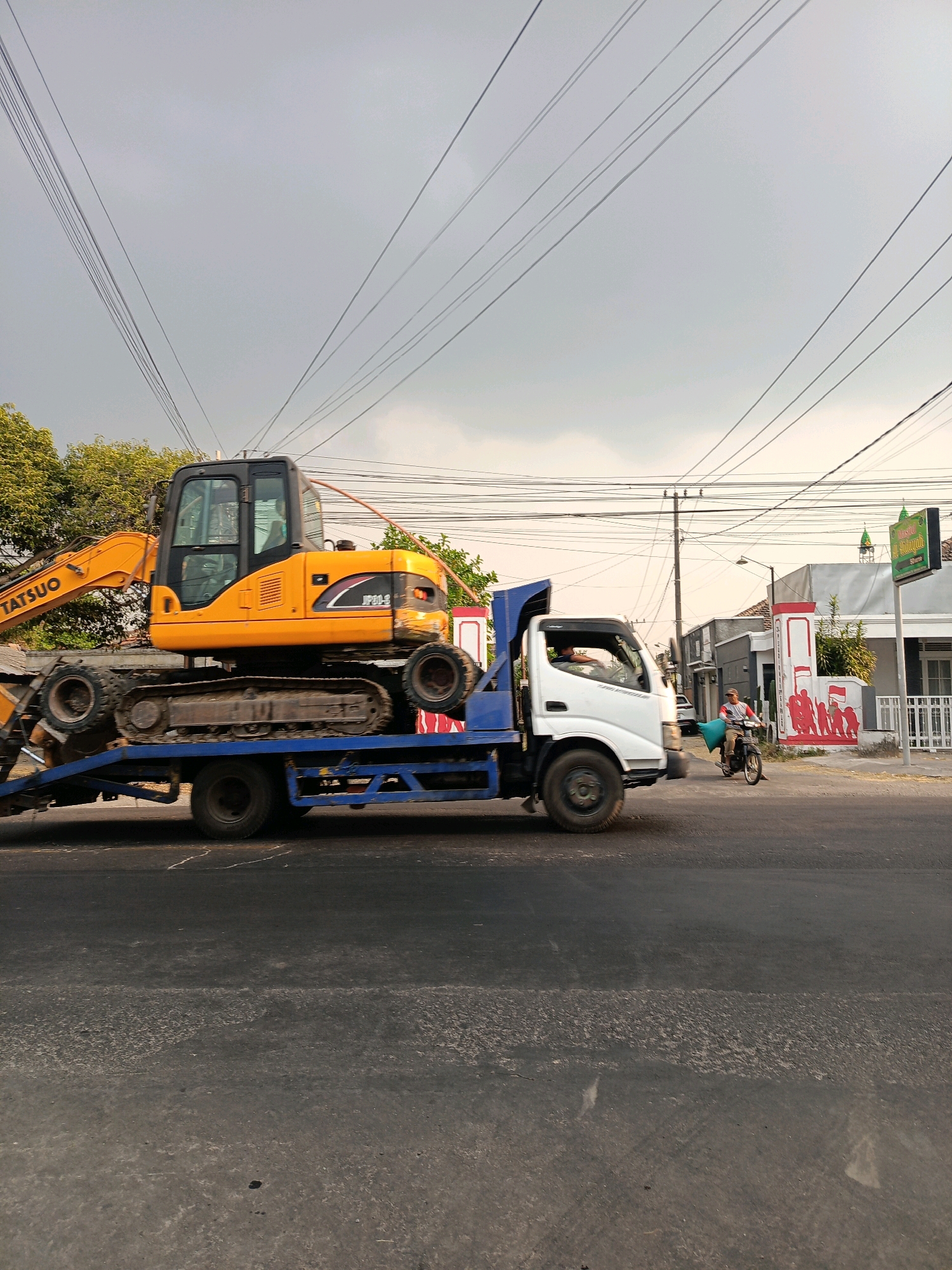 The image depicts a street scene in Kincang Wetan, Indonesia. A flatbed truck, carrying a yellow excavator, is traveling on a paved road. The excavator is positioned horizontally on the truck's bed. The truck is white with a blue bed. The excavator is labeled "Tatsuo" and "JP20-S". In the background, there are buildings, trees, and a cloudy sky with overhead power lines. A person on a motorcycle is visible in the right side of the image, riding past a building with a mural on its wall. The mural appears to depict a group of figures. A green sign with writing in a foreign language is mounted on a pole. The road surface shows signs of wear and tear.