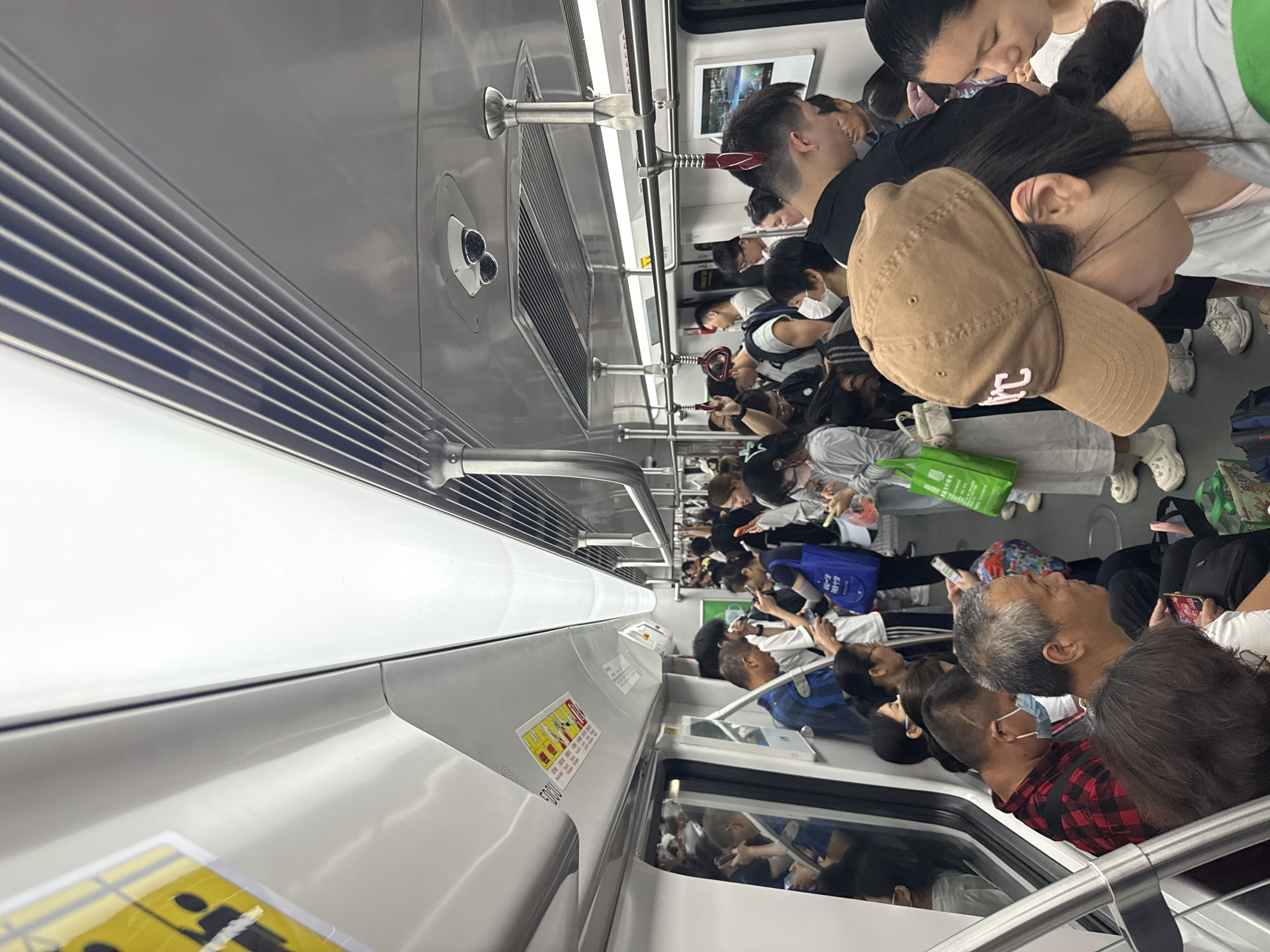 The image depicts the interior of a crowded subway car in Tianhe District, China, captured from a low, upward-looking angle along the side of the train. The scene is densely packed with commuters, primarily adults, standing and filling the available space.
Numerous individuals of various ages are visible, most of whom are standing close together. Many passengers are engaged with their mobile phones, looking down at screens or holding them up. Others are simply standing or looking around, appearing to be in transit. Several people are wearing face masks, and some are carrying bags, including a bright green reusable shopping bag and a blue tote bag. The general atmosphere is typical of a busy urban commute, characterized by a high density of people but a relatively calm demeanor.
The subway car's interior appears modern and clean, with light-colored (likely white or light gray) walls and ceiling, complemented by silver-colored fixtures such as overhead grab handles and railings. A horizontal grille-like ventilation system runs along the upper part of the wall. The lighting within the car is artificial, suggesting it is either nighttime or the train is underground.
Notable details include:
* A yellow rectangular sticker on the side wall features pictograms that appear to convey safety or behavioral instructions, along with some Chinese characters. The number "5001" is clearly visible below this sticker, likely identifying the specific train car.
* In the foreground, a person is seen wearing a brown baseball cap with a light pink, stylized letter or character on its front.
* Other signs with Chinese text are present but are not legible in detail.
* The clothing worn by passengers is casual and varied, reflecting everyday attire.
The image captures a common sight in metropolitan areas, showcasing the heavy usage of public transportation during what appears to be a peak travel time.