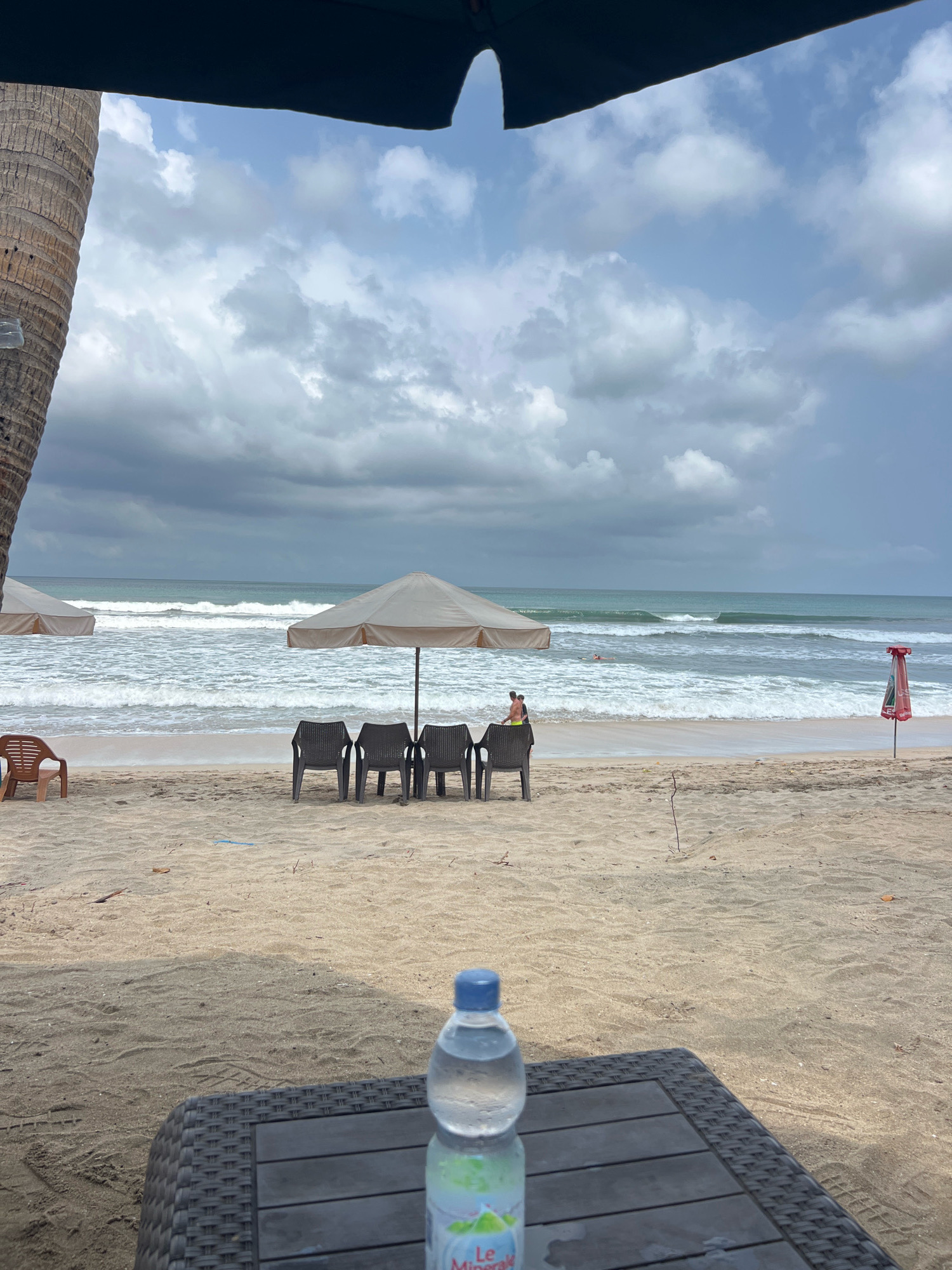 The image depicts a beach scene in Kuta, Indonesia, likely on a cloudy day given the overcast sky. In the foreground, a woven black table holds a clear plastic bottle of water with a blue cap. Beyond the table, the sandy beach stretches towards the ocean. Several dark plastic chairs are arranged in front of a beige umbrella. To the left of these chairs, a single brown plastic chair stands alone. Further back, another beige umbrella is visible, with the ocean and its waves in the background. Two individuals are present near the water, one appearing to be in the ocean, and another on the shore wearing yellow shorts. A red and white umbrella is planted in the sand on the far right of the image, near the water's edge. A large palm tree trunk is partially visible on the left side of the frame.