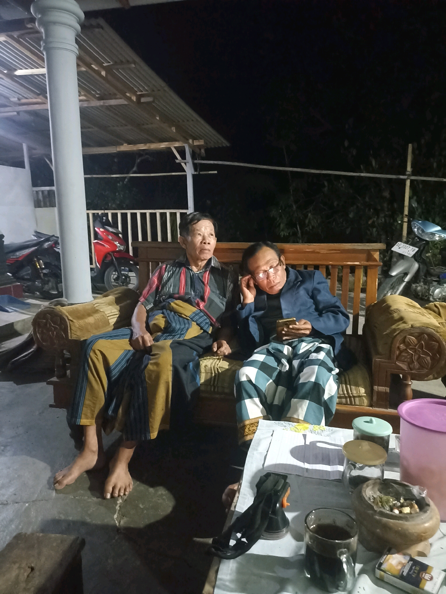 Two adult males are seated on a wooden bench. The male on the left, wearing a striped short-sleeved shirt and a sarong, is looking upwards. The male on the right, wearing glasses and a blue jacket over a dark shirt, is looking at his smartphone, with his left hand resting on his ear. Both men appear to be of Asian descent and are wearing traditional Indonesian clothing.
The bench is positioned outdoors, likely on a porch or patio, as indicated by the presence of a concrete floor and a corrugated metal roof supported by wooden beams. A large white pillar is visible to the left of the frame, and a wooden railing is behind the bench.
In the background, two motorcycles are parked: a red motorcycle on the left and a lighter-colored motorcycle on the right. The environment is dark, suggesting it is nighttime.
In the foreground, a table is set with several items. These include a glass of dark liquid, a pair of black shoes, a cigarette pack, and several containers, one of which appears to be an ashtray with smoking residue. A pink plastic container is also visible to the right of the table. The overall scene suggests a relaxed, informal gathering.
The specific location in the city of Wonogondo, Indonesia, is not visually discernible from the image alone.