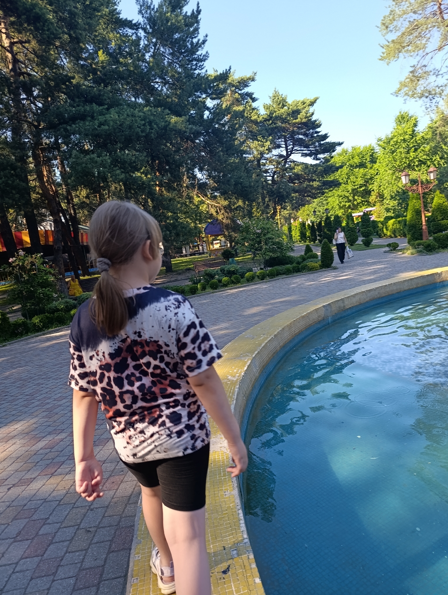 A young girl with blonde hair in a ponytail, wearing a leopard-print t-shirt and black shorts, is standing at the edge of a bright blue fountain. The fountain has a yellow tiled rim. The girl is facing away from the camera, looking towards a park. The park features well-maintained landscaping with manicured bushes and trees. A woman in a white shirt and black pants is walking on a paved pathway in the background, carrying a white bag. The scene is outdoors on a sunny day, with tall trees framing the background and a clear blue sky above. The ground is paved with interlocking bricks.