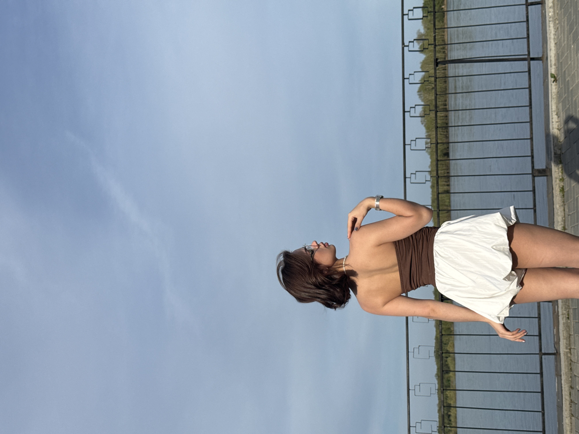 A young woman stands on a concrete walkway beside a metal railing overlooking a body of water. In the background, trees line the opposite shore. The sky is light blue with minimal clouds. The woman wears a brown top, a white skirt, glasses, and a watch. She is facing away from the camera, looking towards the water. The image may be taken in Sarapul, Russia.