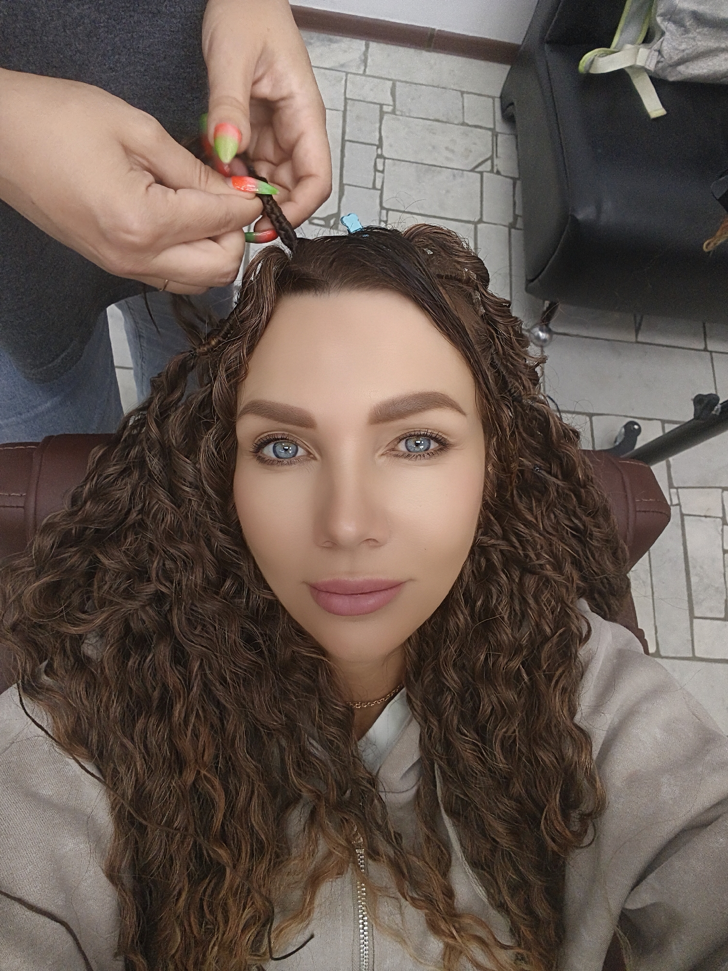 In a close-up, elevated view, a woman with prominent blue eyes and long, medium-brown curly hair looks directly at the camera with a calm, slightly smiling expression. She is seated in what appears to be a brown leather salon chair. Her hair is being styled, with several sections already braided into tight, small plaits, and other sections held back with small blue clips on top of her head.
The hands of another person are visible in the upper part of the frame, actively braiding a section of the woman's hair. These hands have long fingernails painted in a vibrant neon green and orange, and the person wears a grey top and blue jeans.
The woman is wearing a light-colored (possibly beige or grey) hooded top or jacket with a visible zipper and a delicate gold chain necklace.
In the background, to the right, a dark-colored piece of furniture, possibly an armchair, is visible. The floor is covered with light-colored square tiles, suggesting an indoor setting like a hair salon or a home styling area. The lighting appears to be artificial, typical of an indoor environment. No text is visible in the image.