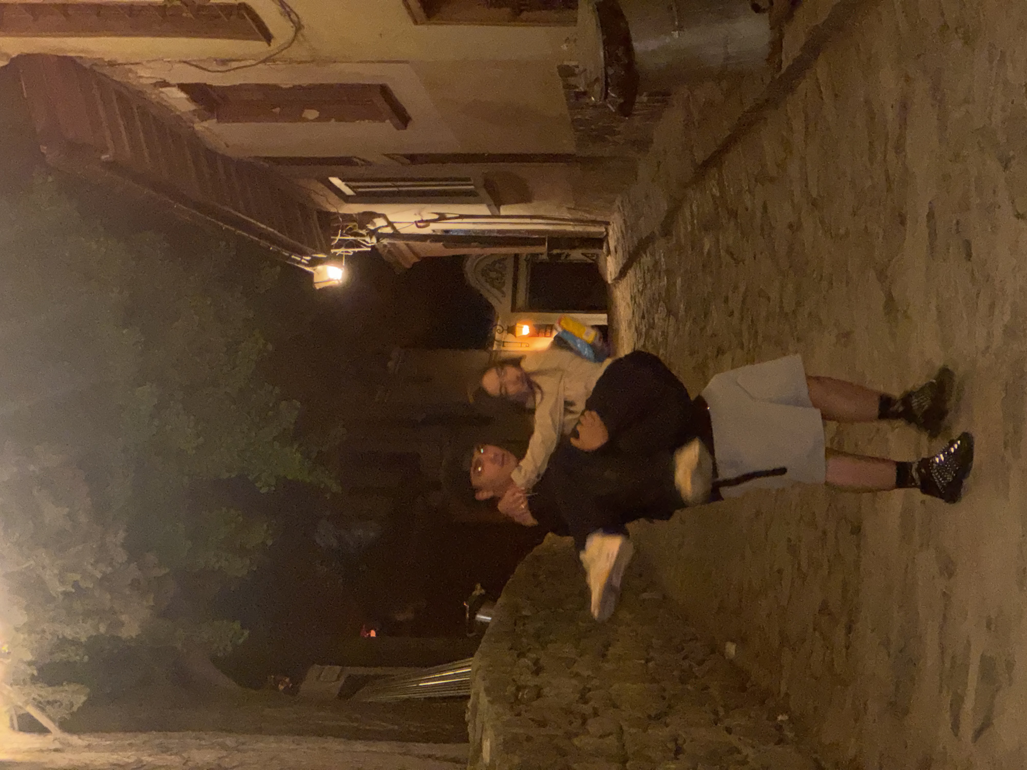 Two people are present in the photograph. One individual carries the other piggyback style. The individual being carried has long hair and is wearing a light-colored top. The person giving the piggyback ride is wearing dark clothing, glasses, shorts and dark shoes. The scene appears to be an alleyway at night with a cobblestone surface. Buildings are visible on either side of the alley, along with some vegetation. The general location resembles a street in the city of Plovdiv, Bulgaria.