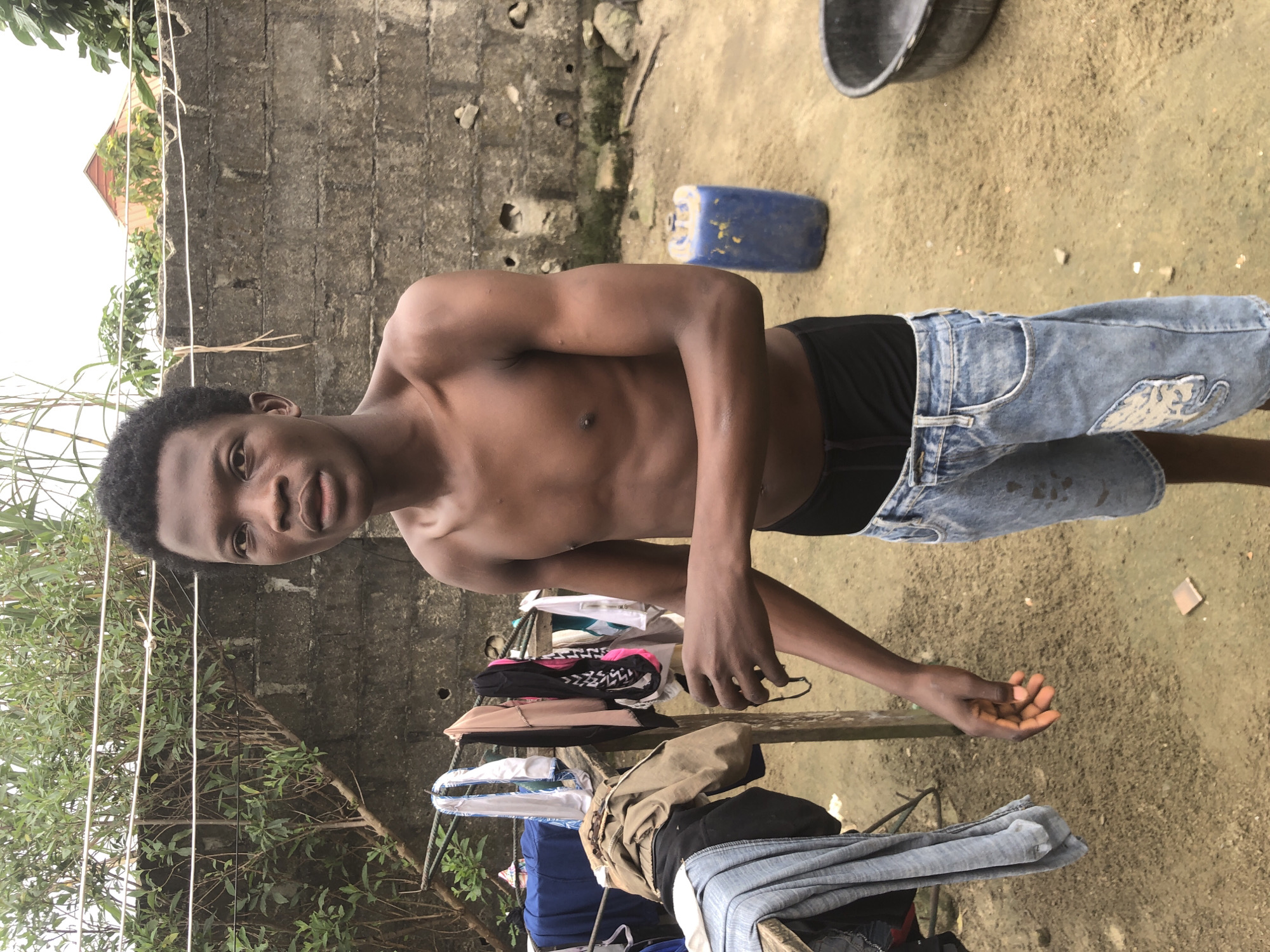 In Ajah, Nigeria, a young Black male stands outdoors in a yard, shirtless, wearing denim shorts with a black waistband. He has short, dark, curly hair and is looking towards the camera with a neutral expression. To his left, laundry is hung on a wooden structure, with various garments, including a blue shirt, a pink and black patterned garment, and a beige garment. Behind him, a textured concrete wall forms the backdrop. A large blue plastic drum and a dark basin are visible on the sandy ground to his right. The environment appears to be a residential backyard.