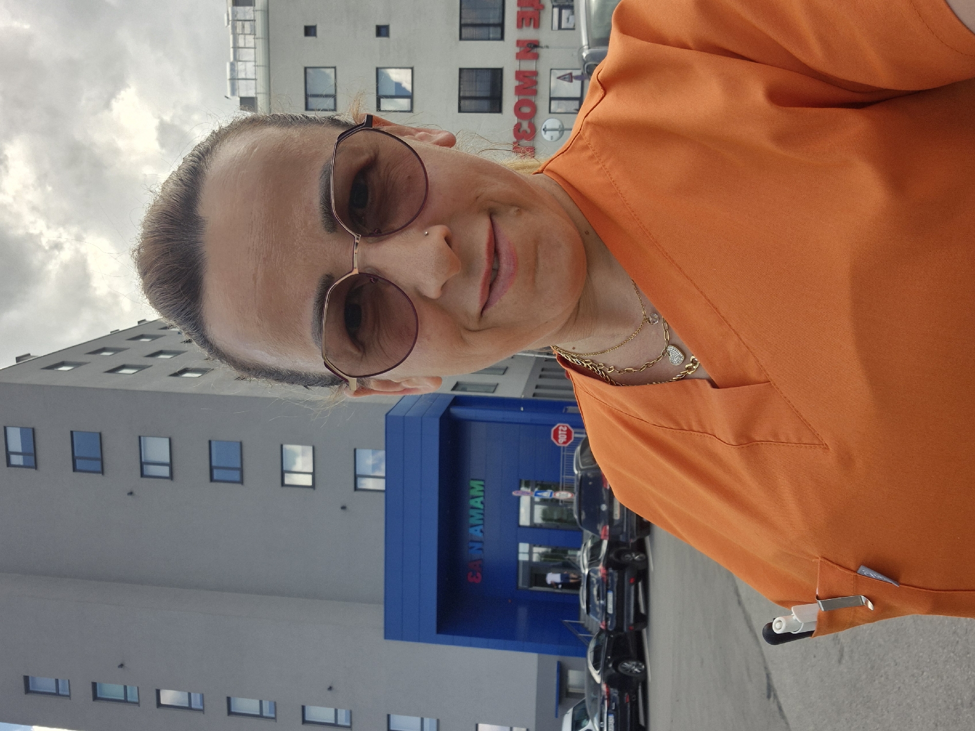 This media file shows a woman in an orange scrub top and large sunglasses posing for a selfie outdoors. She has her hair pulled back and is wearing a delicate gold necklace and a nose ring. Her expression is pleasant, with a slight smile. Behind her, a modern, light-colored building with many windows is visible. To the right, a blue entrance to a building is adorned with green and red lettering that reads "SANAMAM". A red stop sign is also present near the entrance. Several dark-colored cars are parked in a lot in front of the building. The sky is cloudy, suggesting it might be an overcast day. The image is taken from a slightly upward angle, with the building and cars appearing to be on a lower level. The overall scene appears to be a daytime shot in an urban or suburban environment, possibly near a medical facility or office building given the woman's attire and the building's signage.