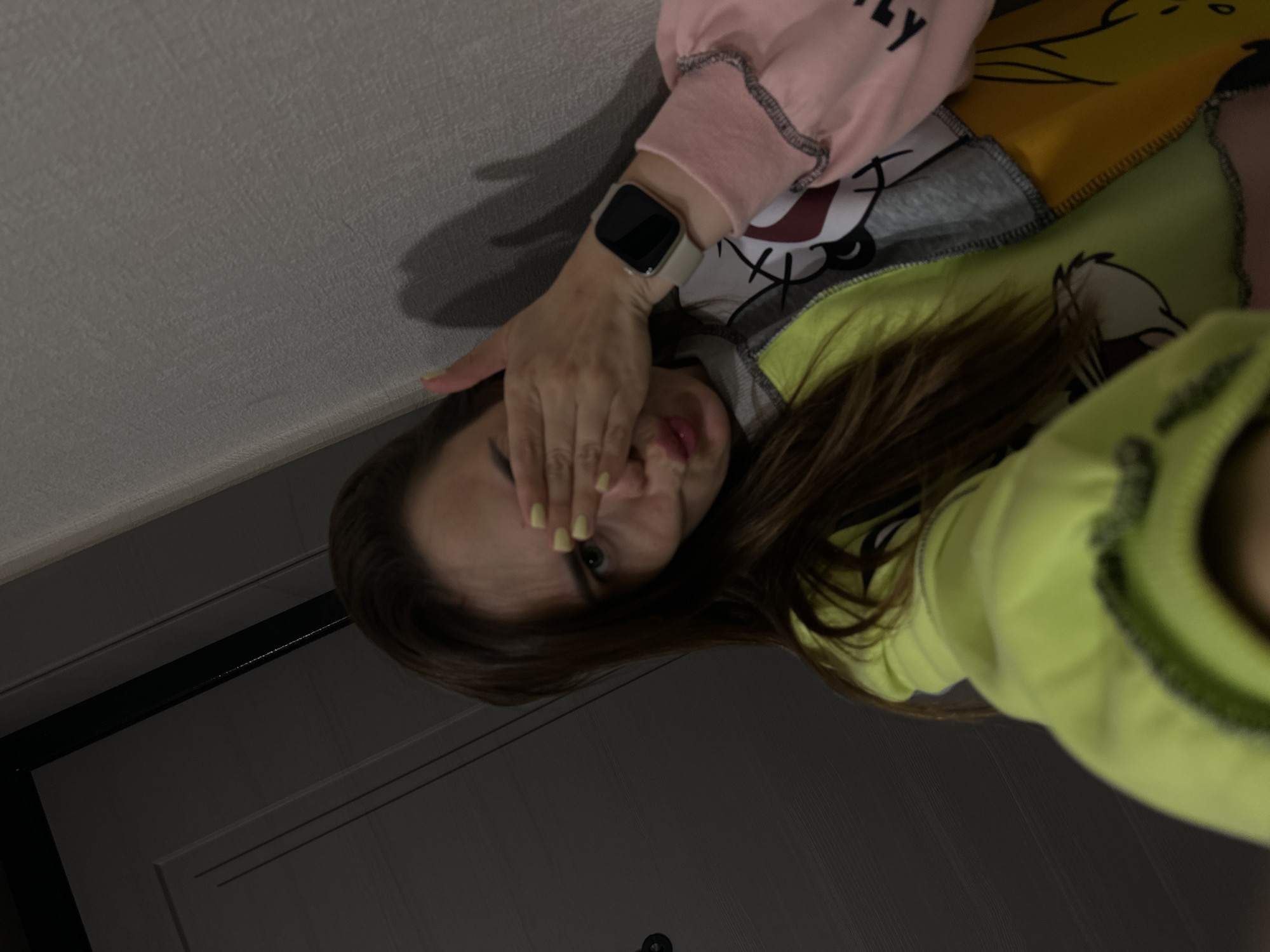 A young woman is taking a selfie in an indoor setting, likely a hallway. She has her right hand covering her right eye, and her lips are pursed. She is wearing a multi-colored sweatshirt with cartoon characters on it, and a smartwatch on her left wrist. The background includes a dark gray door on the left and a textured light gray wall on the right. The lighting is dim, creating shadows. The location of Perm, Russia, is not discernible from the image itself.