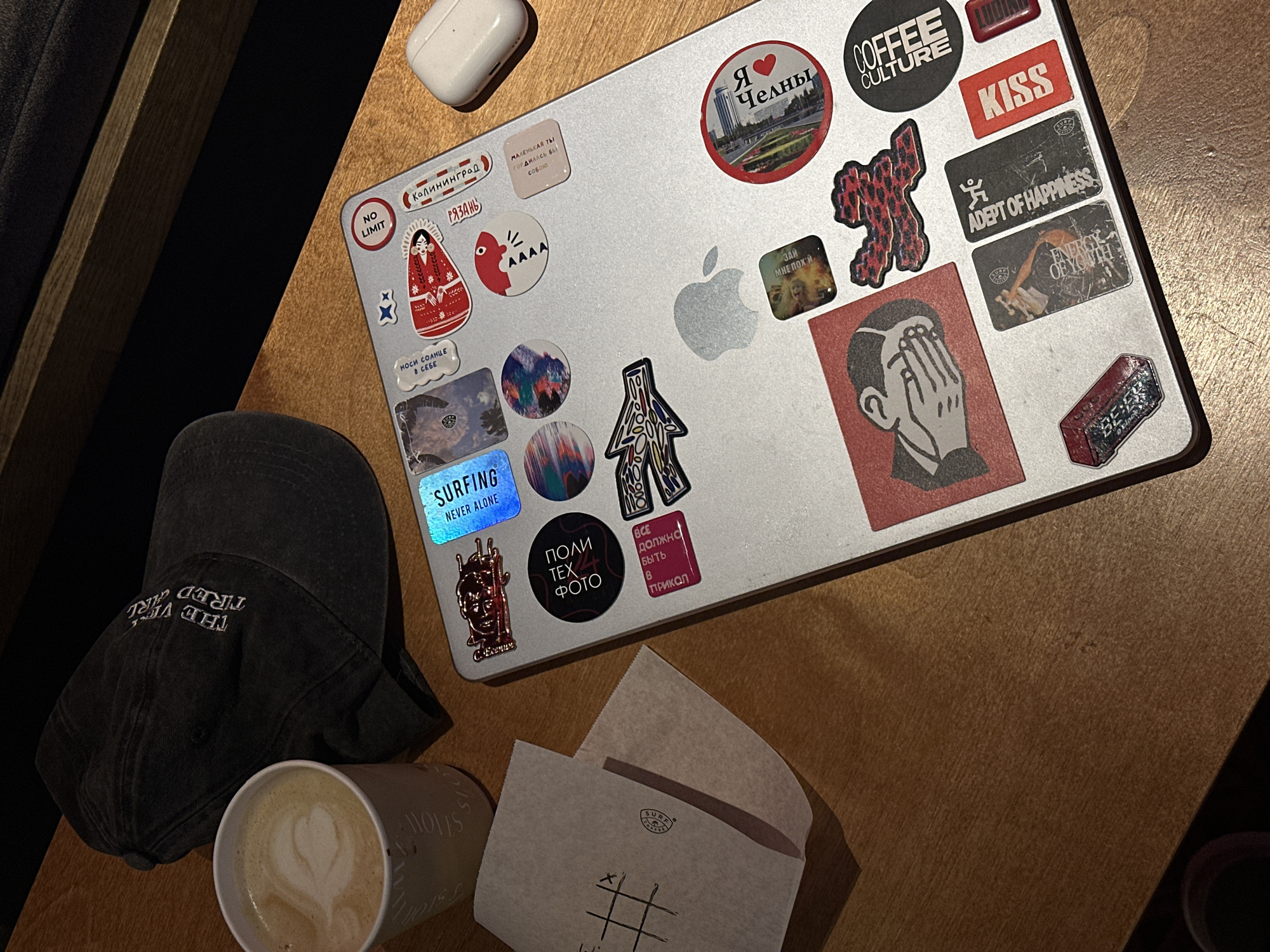 Here is a top-down view of a table containing a laptop covered in stickers, a black cap, a paper cup of latte, and a paper wrapper. The laptop has stickers featuring images and text in both Russian and English. Some of the stickers include "Я Челны", "Coffee Culture", "Kiss", "Adept of Happiness", "Surfing Never Alone", and "Все должно быть 8 прикол". The black cap has "THE VERY TIRED GIRL" embroidered on it. The paper cup contains a latte with a heart-shaped design. The paper wrapper appears to be a napkin with a "SURE" logo and a tic-tac-toe game drawn on it. The table has a wood grain pattern.