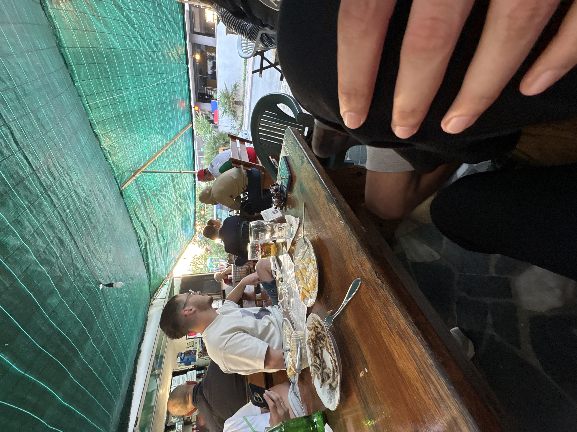 Several people are seated around a long wooden table in an outdoor dining area with a green awning above. They are eating and drinking, with plates of food and beer glasses visible. The location is in Tsarevo, Bulgaria, as evidenced by the Bulgarian flag flying outside.