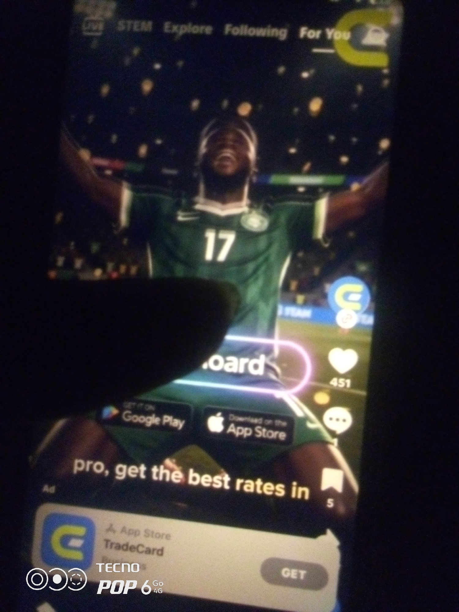 The image is a close-up, slightly blurry photograph taken of a smartphone screen, likely in the city of Howayi, Nigeria. The screen displays what appears to be a social media or video sharing application, with various interactive elements. In the foreground, a dark, shadowy hand is obscuring part of the lower left portion of the screen.
The main subject on the screen is a man in a green sports jersey with the number "17" visible on it. He is looking upwards with his mouth open, appearing to be in a celebratory or excited pose. Behind him, the background is dark with scattered yellow and white lights, resembling a stadium or an outdoor night scene.
Overlaying the video content are several UI elements common to such platforms. At the top, there's a navigation bar with text labels such as "LIVE," "STEM," "Explore," "Following," and "For You." To the right of "For You" is a circular icon with a yellow "C" inside. Along the right edge of the screen, there are vertical icons for likes (a heart with "451" below it), comments (a speech bubble icon with "1" below it), and a bookmark icon with "5" below it.
Below the man's image, there is a prominent translucent pink circle with text inside, partially obscured but appearing to say "board." Further down, there are buttons to "GET IT ON Google Play" and "Download on the App Store." Below these app store links, a large text advertisement reads "pro, get the best rates in". At the bottom of the screen, there's an ad banner with a "C" logo, "App Store," and "TradeCard" visible. Below that, the branding "TECNO POP 6 Go 4G" is displayed.
The overall scene is a digital advertisement or promotional content displayed on a mobile device, with the physical presence of a hand and the context of the device being used. The lighting suggests the photograph was taken in a dimly lit environment.