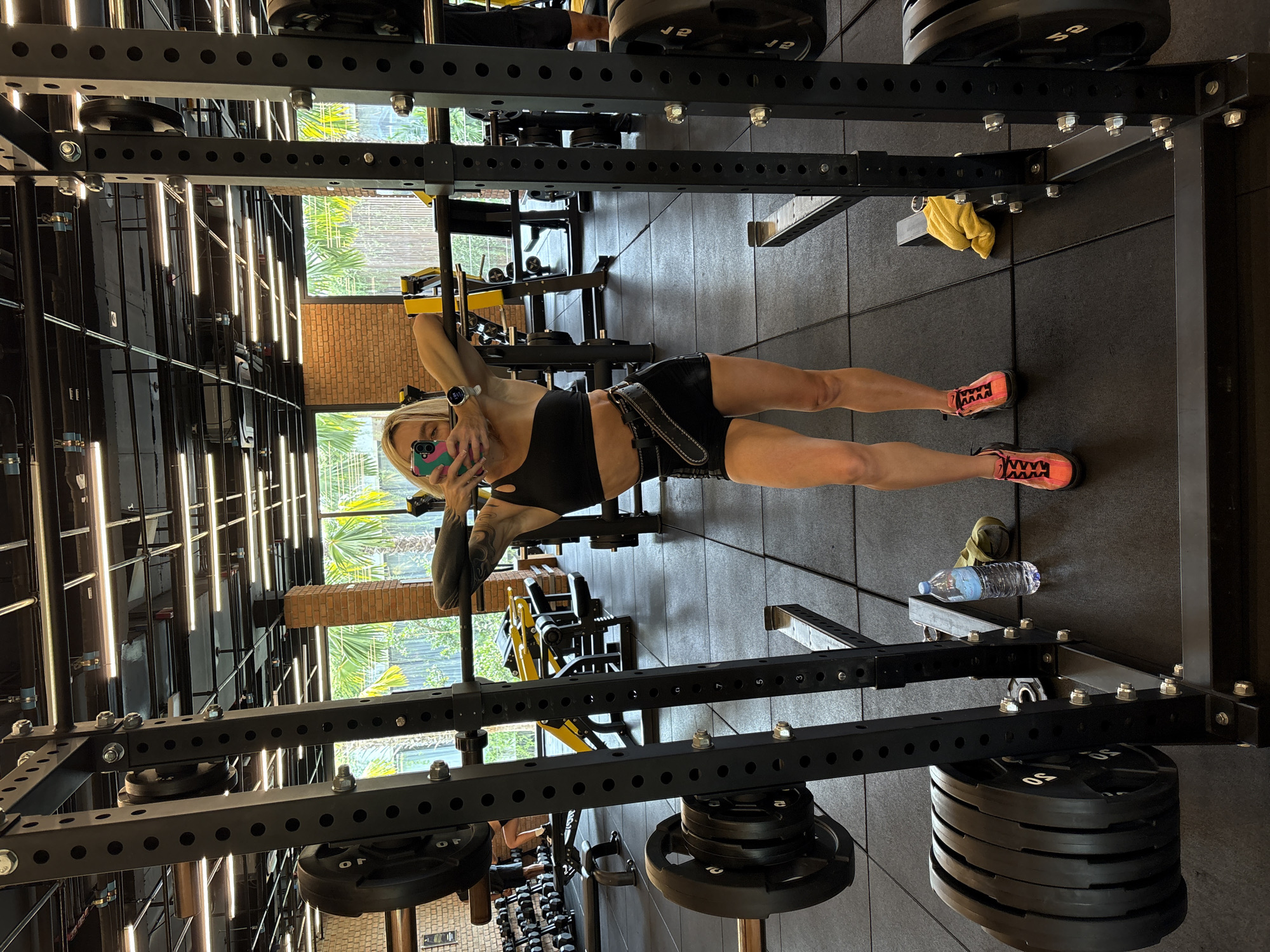 A blonde woman in athletic wear is taking a selfie in a gym. She is standing in front of a squat rack, holding her phone with her right hand. She is wearing a black one-shoulder sports bra, black shorts, a black weightlifting belt, and bright orange athletic shoes. The gym has a dark rubber floor and exposed brick walls. In the background, other gym equipment, including weight plates and benches, are visible. Large windows in the back reveal palm trees and lush greenery, indicating a tropical outdoor environment. There is a water bottle and a yellow towel on the floor near the squat rack. The overall lighting suggests it is daytime.
