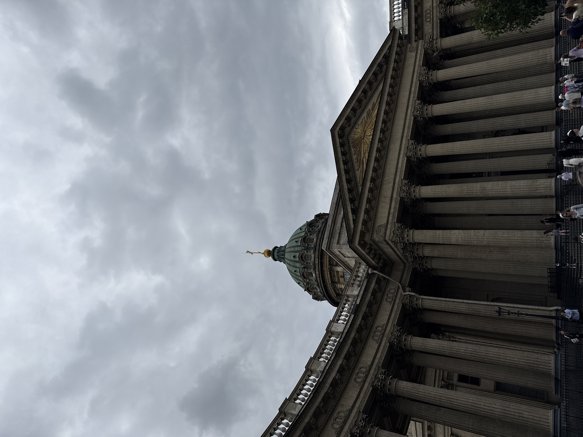 This is a low-angle, full-shot photograph of the Kazan Cathedral in Saint Petersburg, Russia. The sky is overcast with heavy gray clouds. The grand facade of the cathedral is visible, characterized by a semicircular colonnade of Corinthian columns. A large, green dome topped with a golden cross stands prominently in the upper central portion of the image. A triangular pediment with a golden sunburst motif is situated above the central portico. Several people are visible in the foreground, gathered on the steps and walkway in front of the cathedral, appearing to be tourists. The lighting is diffused due to the cloudy weather.