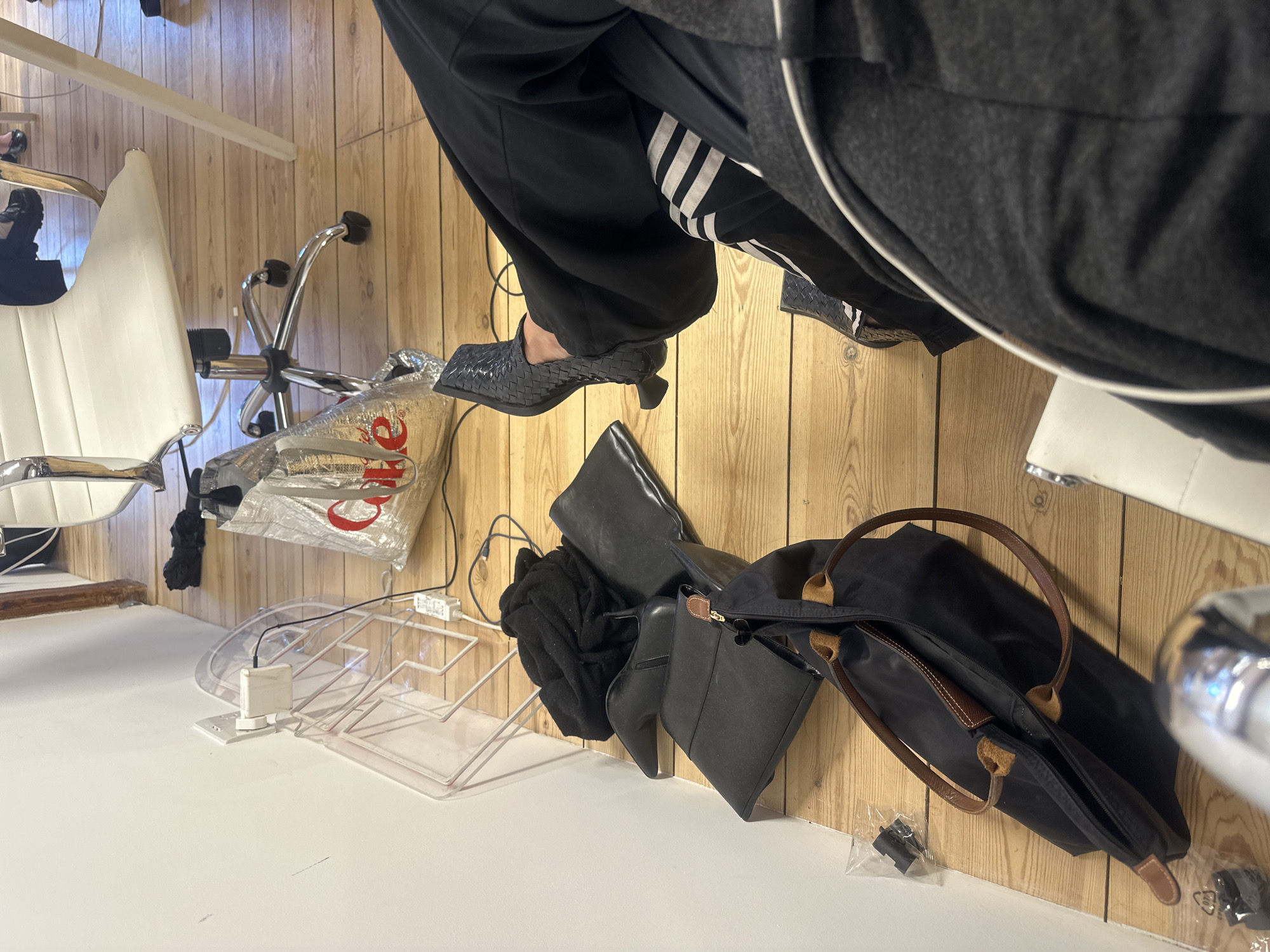 The image shows an interior scene with a mix of personal items and furniture. A person wearing black Adidas track pants and woven shoes is present. Adjacent to the person is a white office chair, a silver Coca-Cola bag, and a transparent plastic shelving unit. Further into the background are multiple black bags, including a Longchamp tote bag with brown leather handles. The environment is characterized by light wood paneling on the walls and a white floor. There is no information about the location, in London, United Kingdom, from the picture.